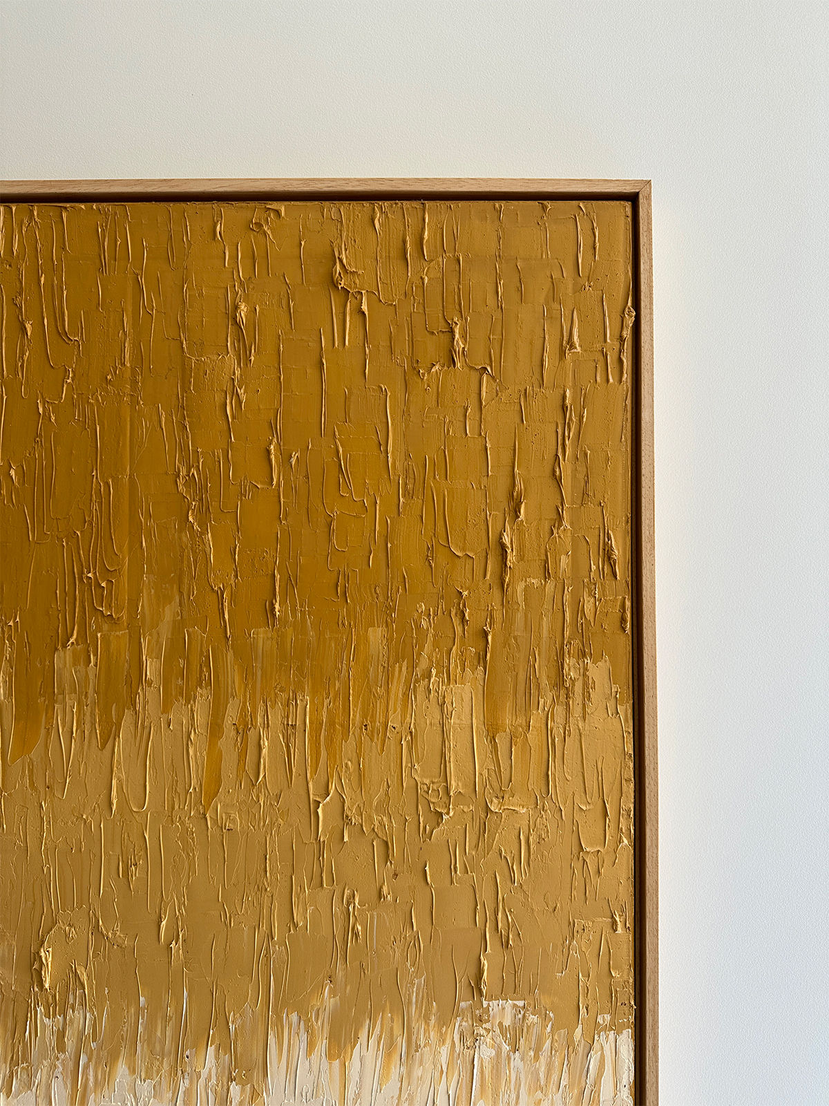 The "Ochre Yellow Faded" oil on canvas by Roi Elmaliah features abstract paintings with textured brushstrokes in shades of gold and brown, forming a layered vertical pattern. The piece is framed with a simple wooden border and displayed against a plain white wall, evoking the minimalist charm of an Amsterdam gallery.