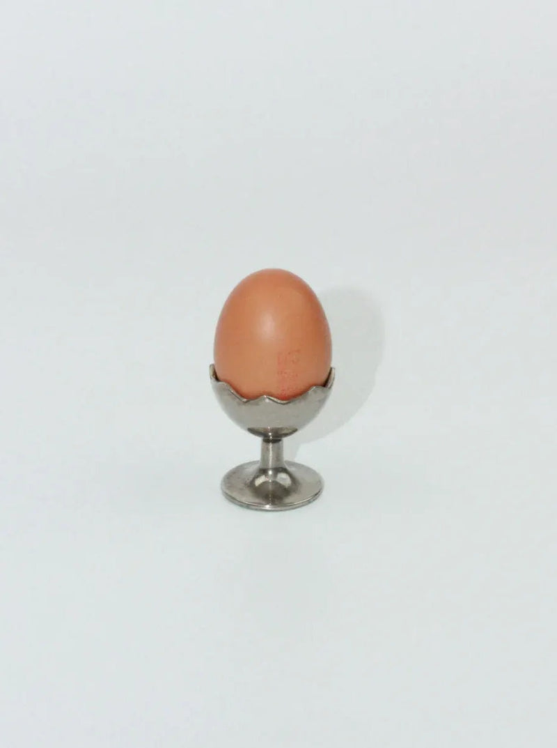 A single brown egg sits upright in a small, silver Boga Avante Shop Egg Cup with a decorative edge against a plain white background. The shadow of the egg and cup is visible to the right.