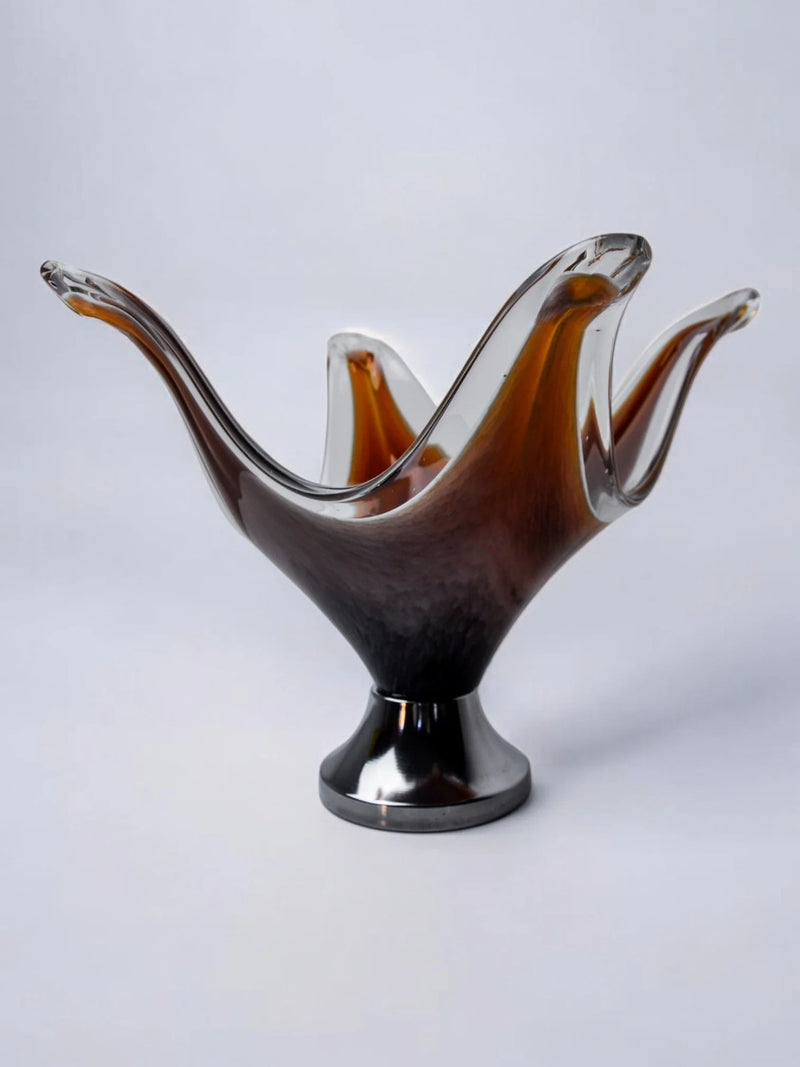 The "Centerpiece by Seguso" from Malamar Studio, crafted in 1970s Murano, Italy, features a decorative translucent glass bowl. Its abstract wave-like design blends amber and black hues using the sommerso technique, resting gracefully on a round base against a plain light gray background.