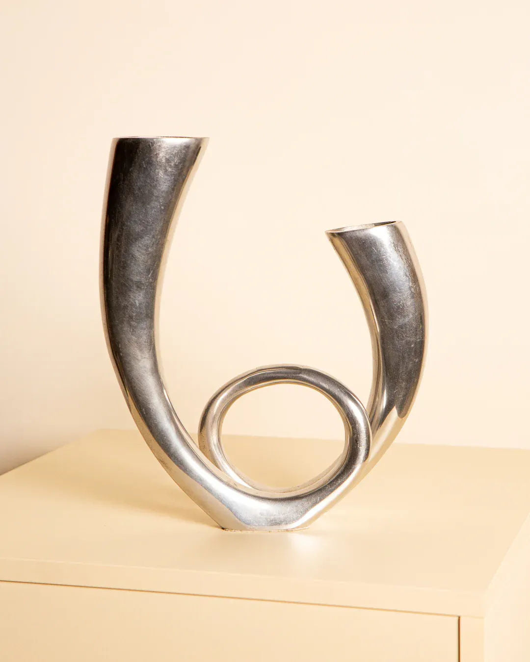 On a beige table stands Treaptyque's 80's Italian Aluminum Double Vase, a modern silver piece with an abstract design. It features two upward-pointing tubes and a circular loop at the base, creating a dynamic shape inspired by 1980s art.