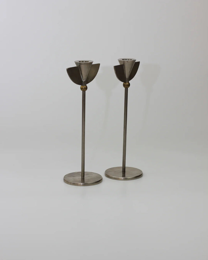 Two tall, sleek stainless steel Memphis Candleholders 80s from Boga Avante Shop stand side by side on a plain background. Each features a circular base and an angular cup at the top, echoing a minimalist, abstract style with hints of Memphis Milano design.