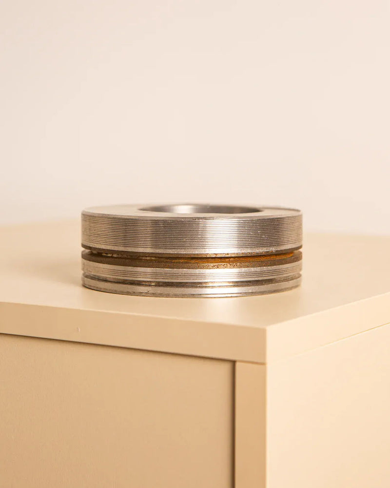 Resting on a beige rectangular surface against a neutral background is the Italian "Piston" ashtray in 80's metal by Treaptyque. This piece, displaying traditional Milanese design, features grooved edges and a circular hole in the center. Its subtle tarnishing enhances its smooth surface with added texture and vintage charm.