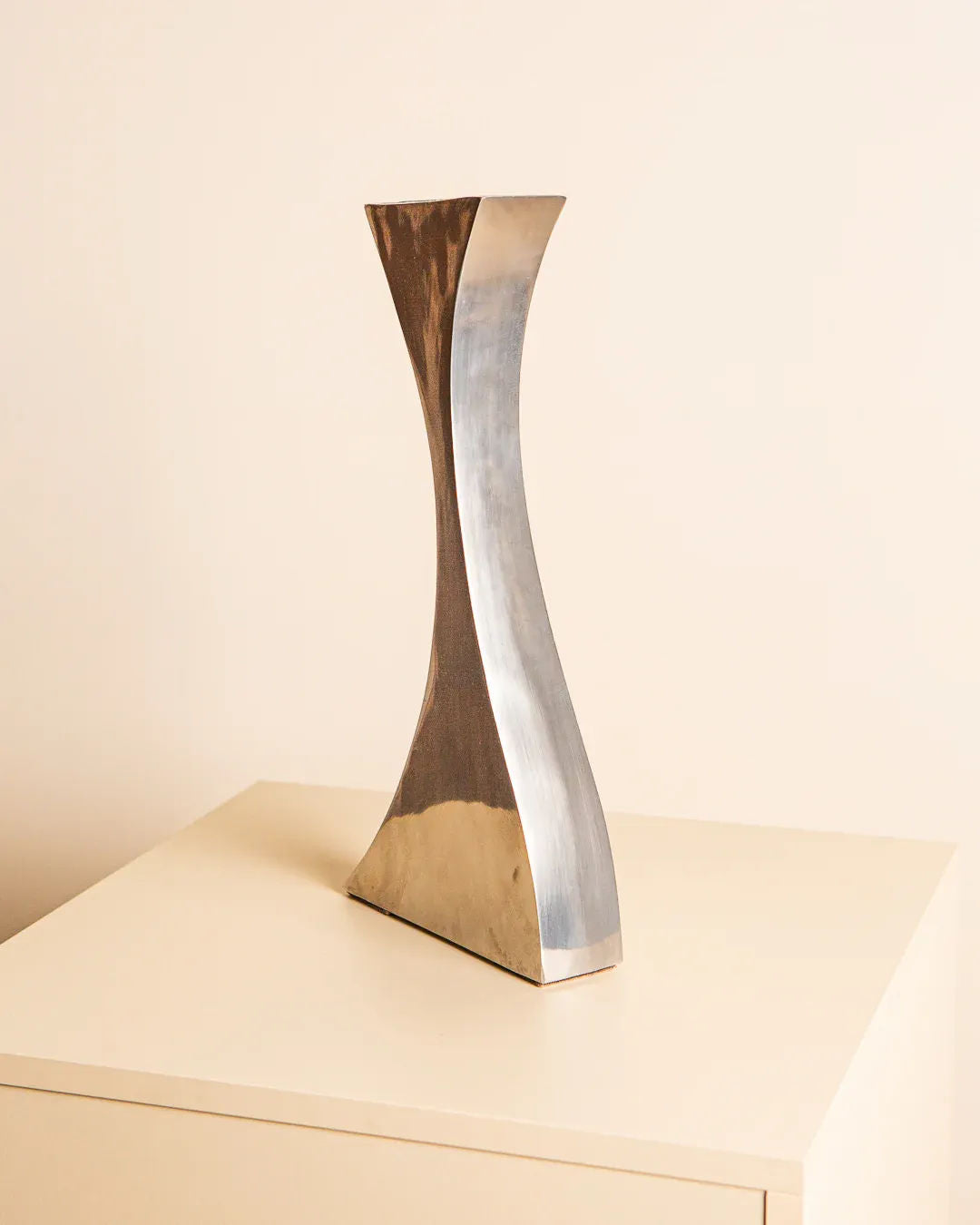 Standing elegantly on a beige, boxy surface against a light-colored wall is the Curved Aluminum Vase 80's by Treaptyque. With its sculptural and twisted design, this vase reflects light beautifully, showcasing its shiny texture and sleek, modern appearance.