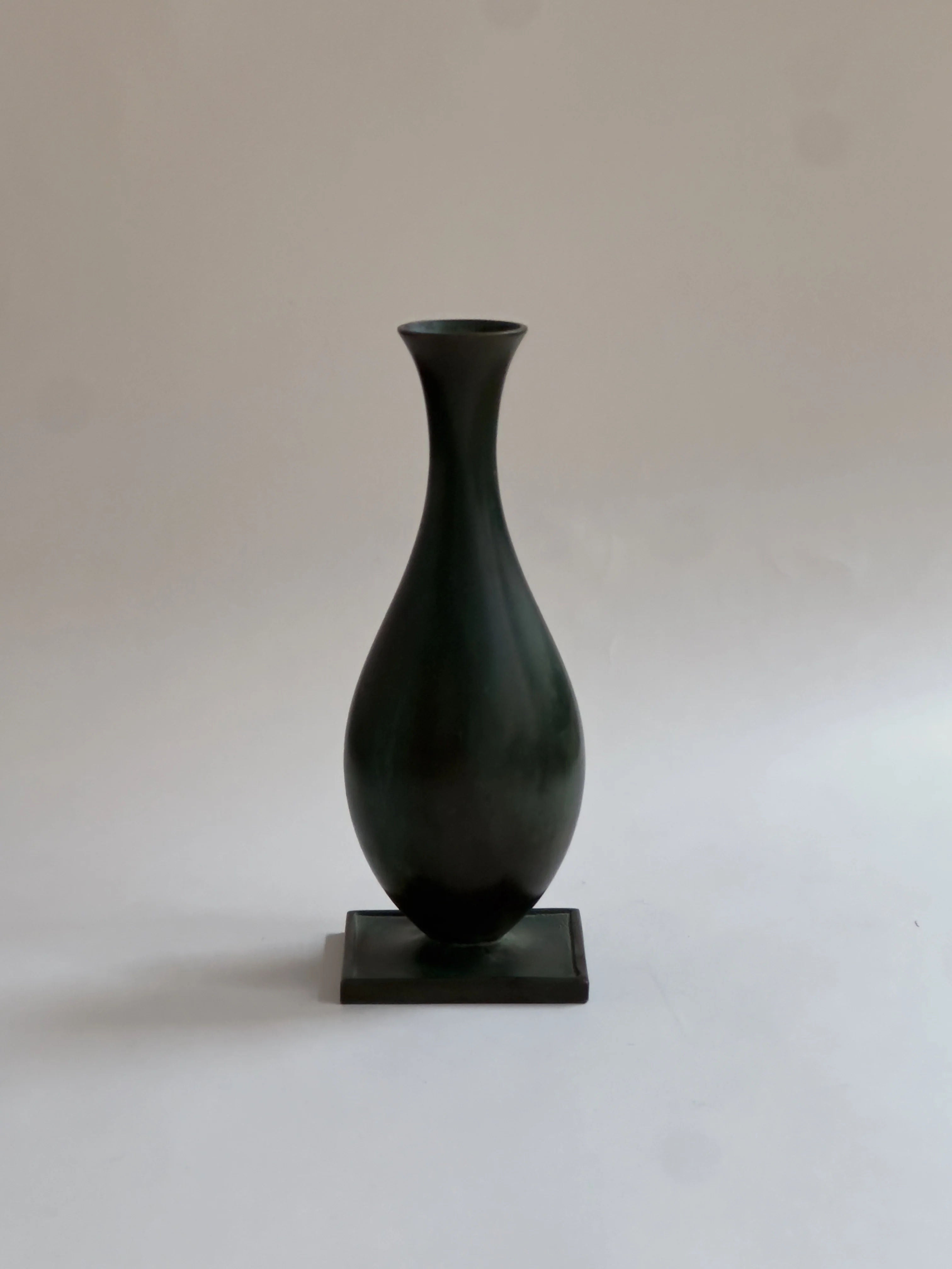 The GAB Bronze vase by Galerie Storm, featuring a sleek black ceramic design with a narrow neck and wide rounded base, stands elegantly on a small rectangular platform. It evokes the elegance of a 1930s patinated bronze vase against a simple light background.