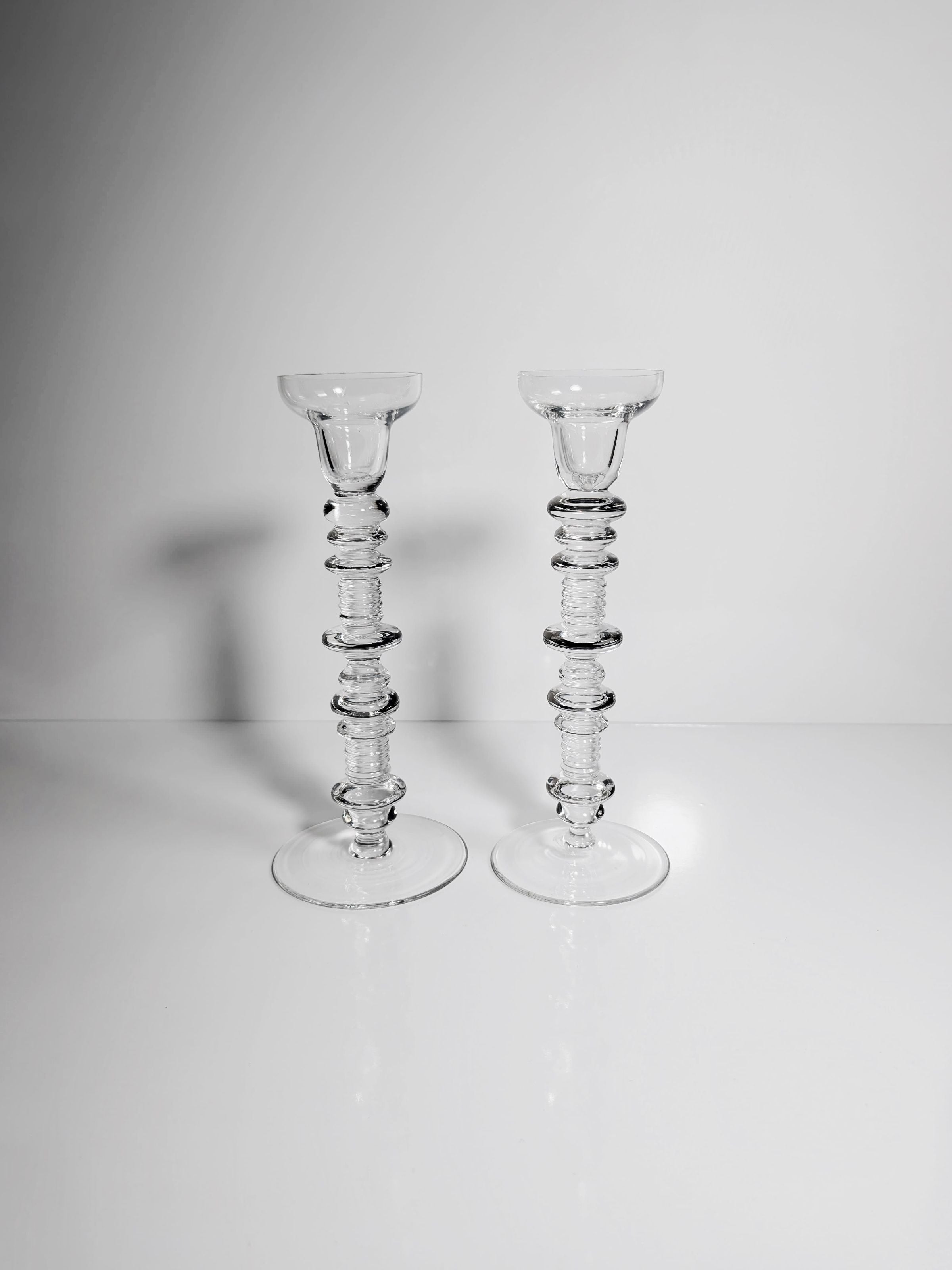 A Set of 2 Glass Candleholders by Peill & Putzler from Dodo Vintage, featuring a textured, beaded design, are elegantly paired on a glossy white surface against a light gray backdrop. The lighting casts delicate shadows that accentuate their transparent mid-century architecture.