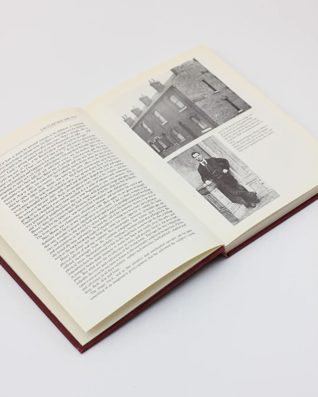 A copy of "Henry Moore, An Illustrated Biography" from Boga Avante Shop rests open on a white surface, displaying a text-filled page on the left and two black-and-white photos on the right. The upper photo portrays a series of brick houses, whereas the lower one showcases a man standing before modern sculpture.