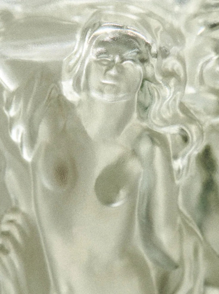 A close-up image of a frosted glass sculpture depicting a partially nude female figure with detailed, flowing hair and an arm raised near her head. The sculpted figure exudes a sense of grace and serenity, reminiscent of Bohemian glassware, with soft, diffused lighting enhancing its delicate features. This beautifully crafted piece is the Michael Vase from Akua Objects.