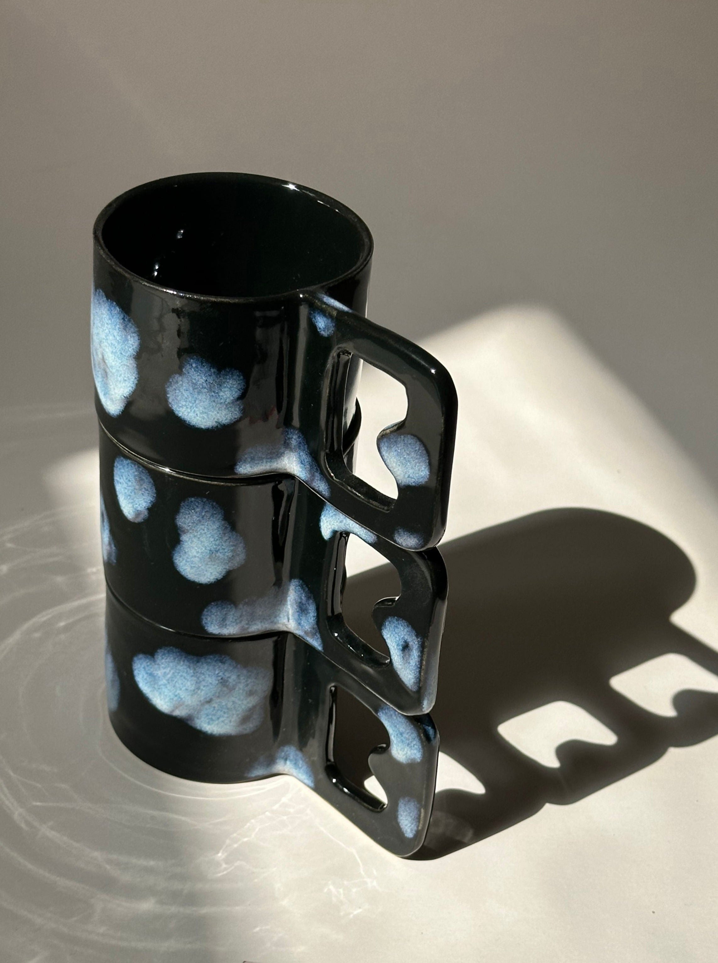 Two black ceramic mugs with blue cloud-like patterns, crafted from stoneware clay and finished with food-safe glazes, are stacked in a vertical alignment. The Cloud Cup by noki ceramics casts long shadows on a light-colored surface, creating a visually intriguing play of light and shadow.