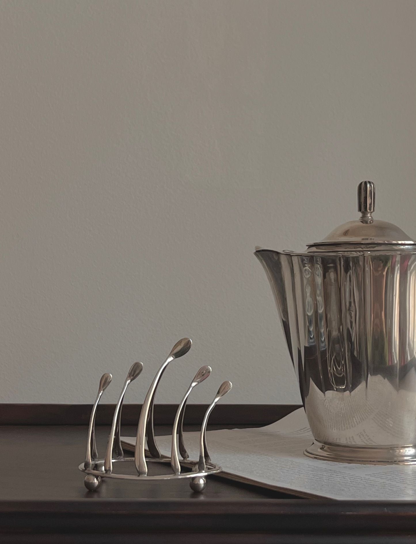 The sleek, silver Wishbone Silver Toastrack by Médecine sits on a dark surface next to a modern napkin holder with spoon-like arches. The plain, light-colored wall in the background highlights the elegant simplicity of this refined setting.