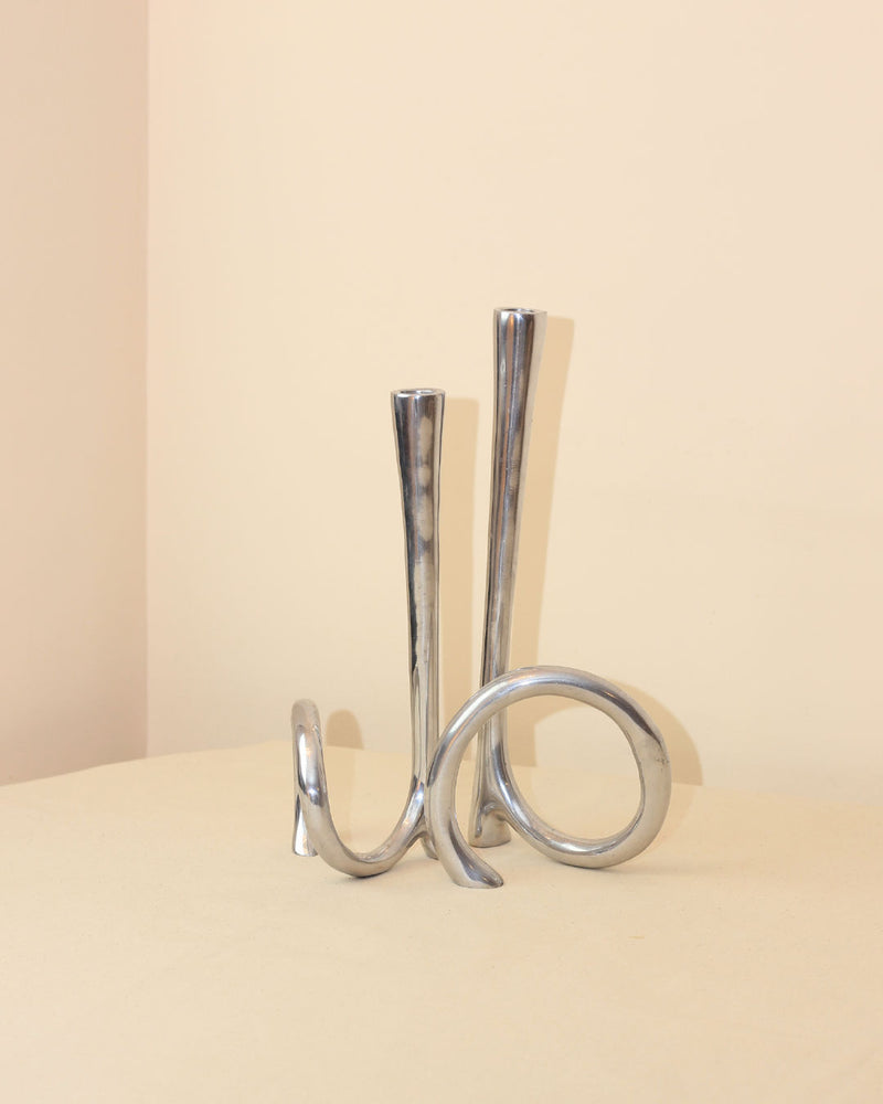 The Boga Avante Shop Cast Aluminum Candleholders feature polished silver metal with two tall, curved stems and an abstract looping base—evoking a modern 1980s candelabra—ideal for minimalist decor on any neutral surface.