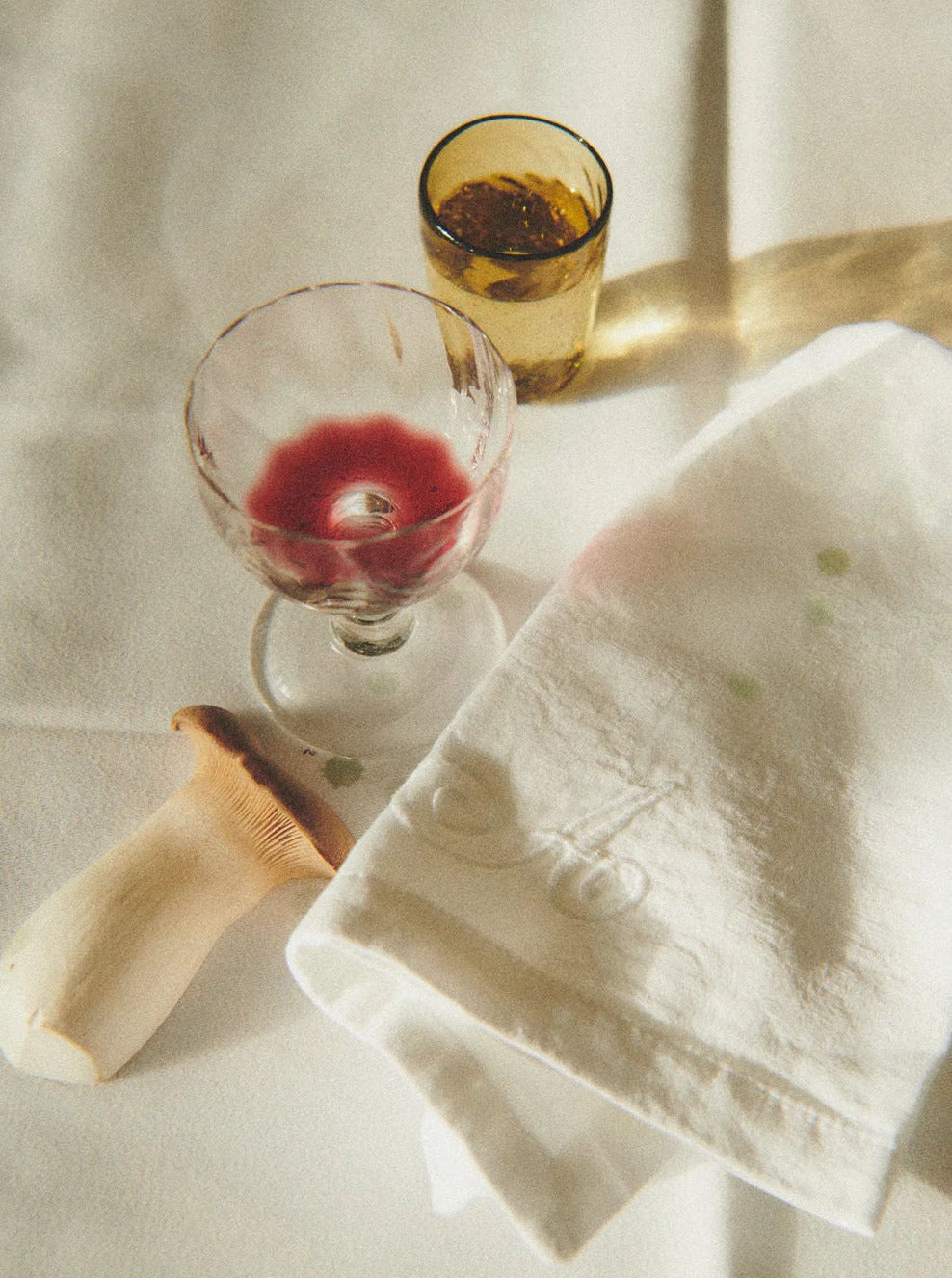 A vintage-style photograph of a table setting with a wine-stained cloth, a half-empty glass of red wine, and another glass from the Akua Objects Alban Wine Glass Small - Set of Four collection with a golden liquid, illuminated by soft, natural