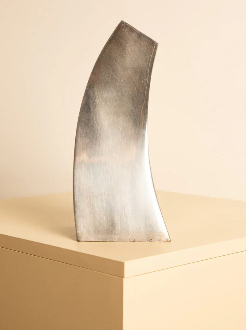 A metallic abstract sculpture with a curved, elongated shape sits on a beige pedestal against a light-colored background. Reminiscent of an '80s metal sculpture, the Treaptyque 80s Metal Vase Sculpture has a smooth surface with subtle reflections, creating a dynamic wave sculpture that embodies a minimalist and modern aesthetic.