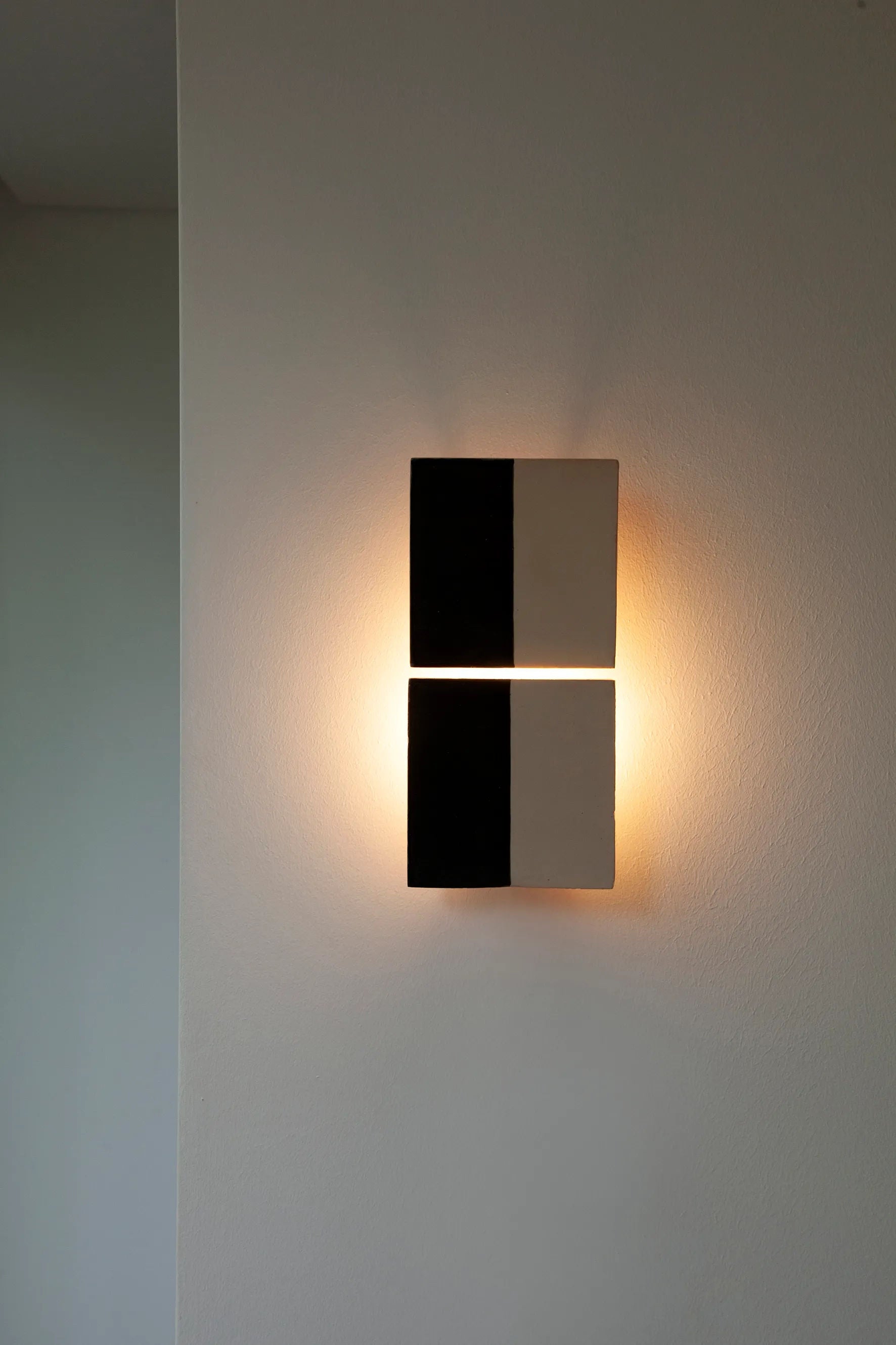 Tiles Line N by Violaine d'Harcourt is a modern wall light fixture with a minimalist design featuring a grid of four rectangles, two black and two white. This handmade, artisanal piece softly illuminates the surrounding wall.