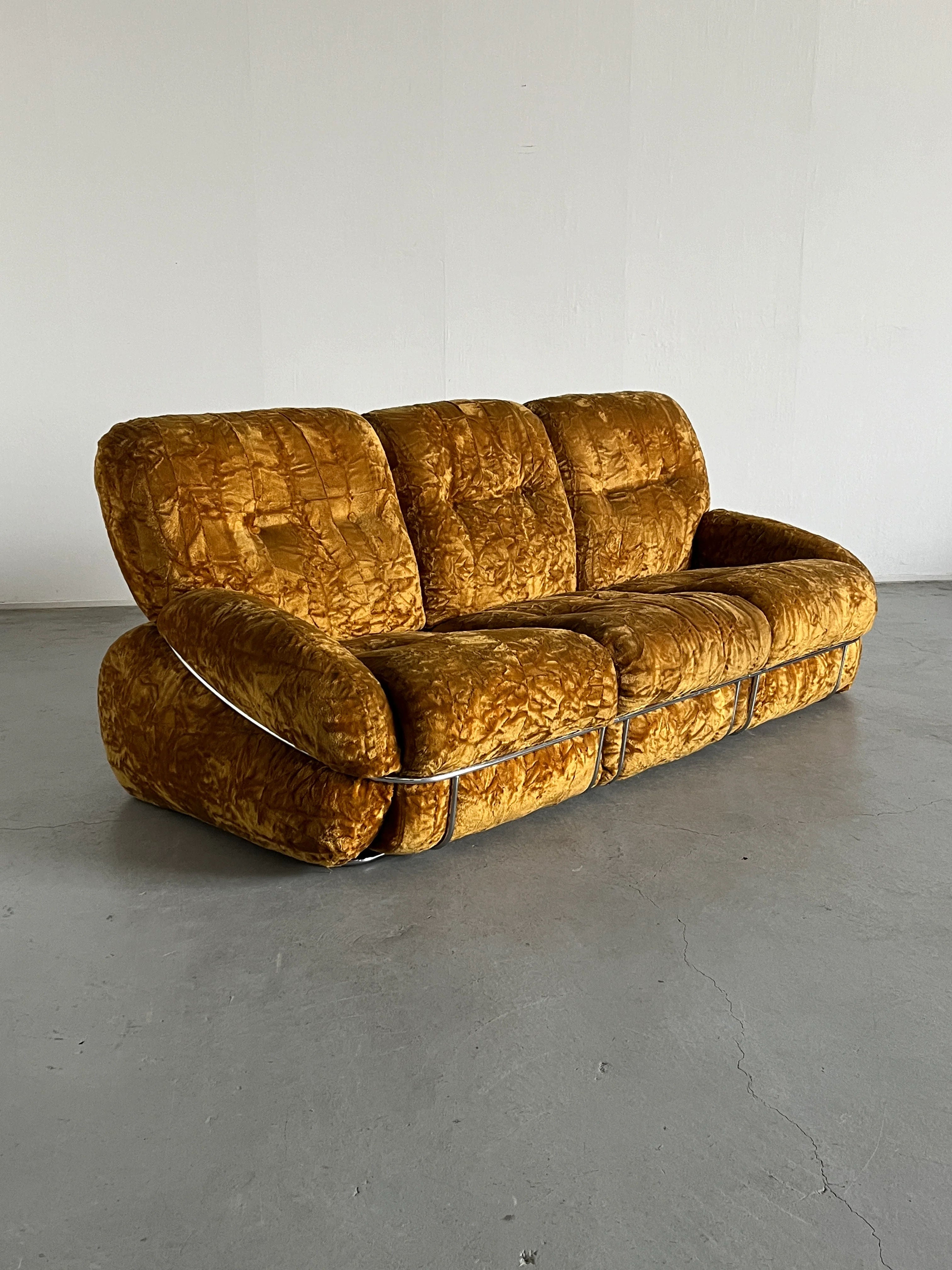 The Adriano Piazzesi 'Okay' Sofa Seating Set from Cherry Cargo, a vintage mustard-colored velvet piece with plush cushions and chrome accents, rests on a gray concrete floor in a minimalist room with white walls.