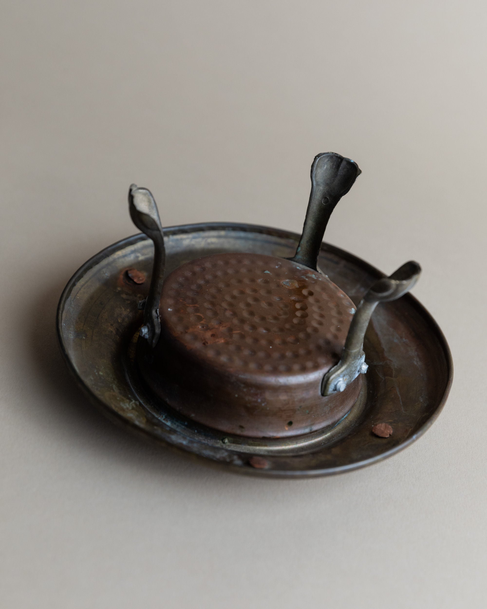 The Vintage Copper Candle Holder by Bottega Jacobs features a brutalist design with three handles and rests on a larger matching plate. Handcrafted from tarnished copper, it boasts a textured surface that enhances its antique charm, all set against a neutral beige background.