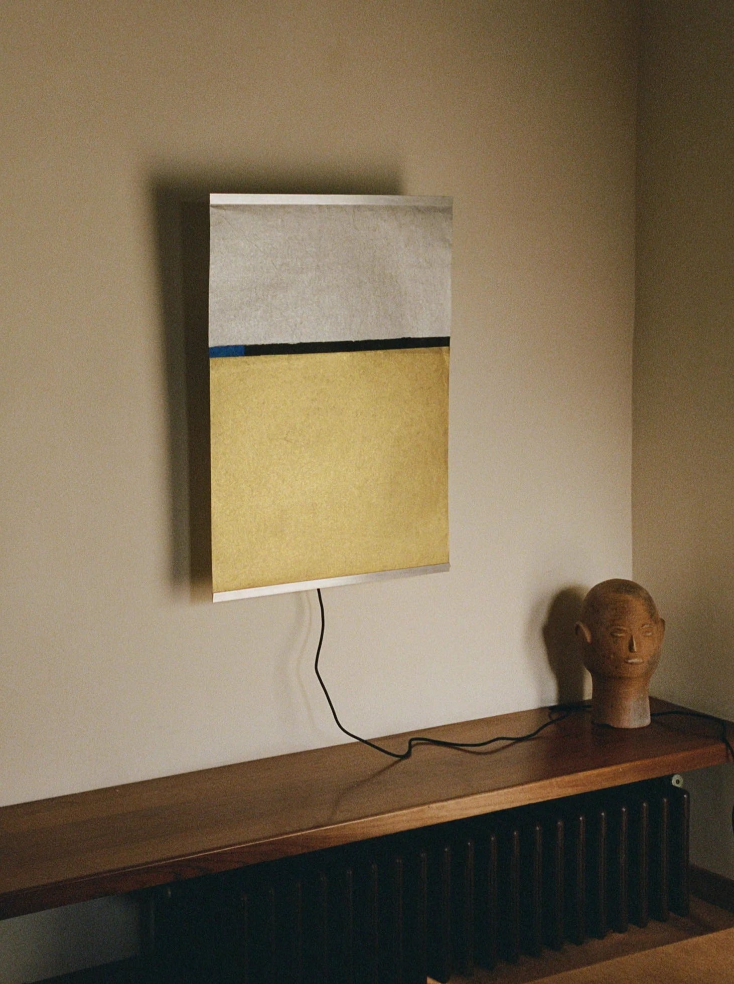 A minimalist artwork featuring sections of white, black, and yellow is mounted on a wall. Positioned below it, a wooden surface displays a handmade stone head sculpture. A cord extends from the TEGET Panel Wall Lamp – Wheat Field attached to the artwork, indicating it might be illuminated by this source.