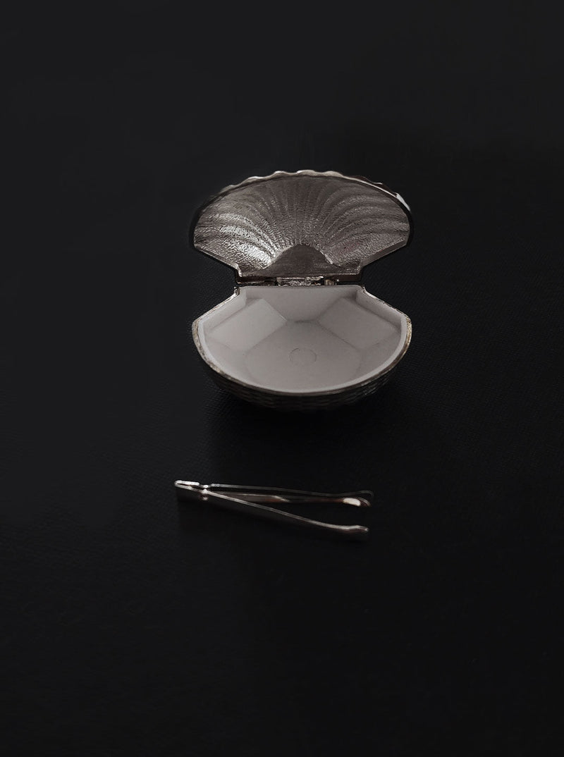 A silver Les Objoies clamshell-shaped pill box opened on a black surface, accompanied by a small metal tweezer to the right.