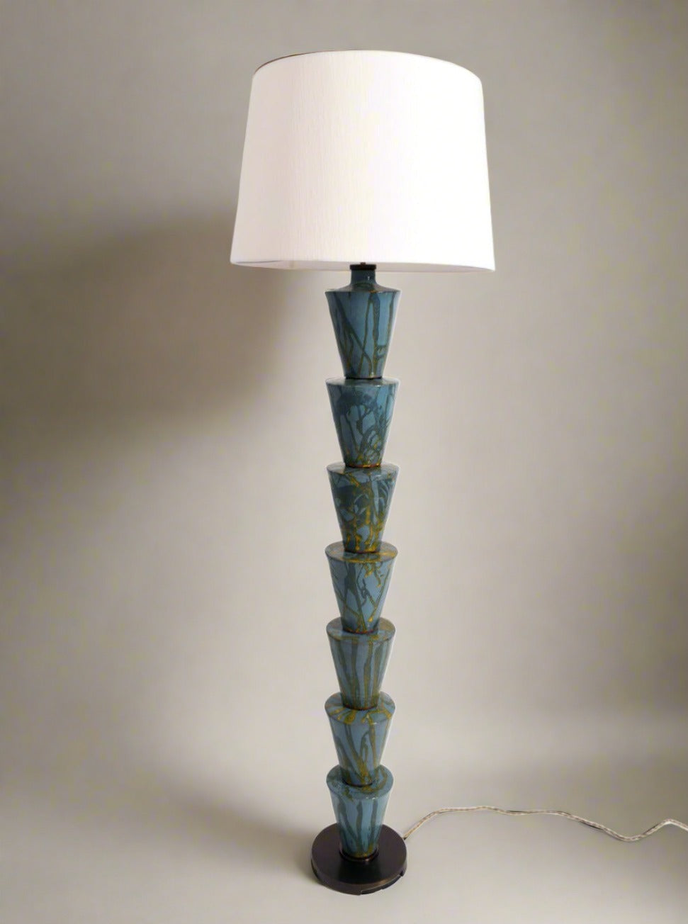 A tall, modern stoneware glazed "Nizwa" Floor Lamp by Barracuda Interiors with a stacked, geometric blue-green ceramic base and a large, white lampshade. The base consists of six cone-shaped sections with gold veining and sits on an iron platform. This exquisite piece, made in Portugal, features a power cord extending from the base.