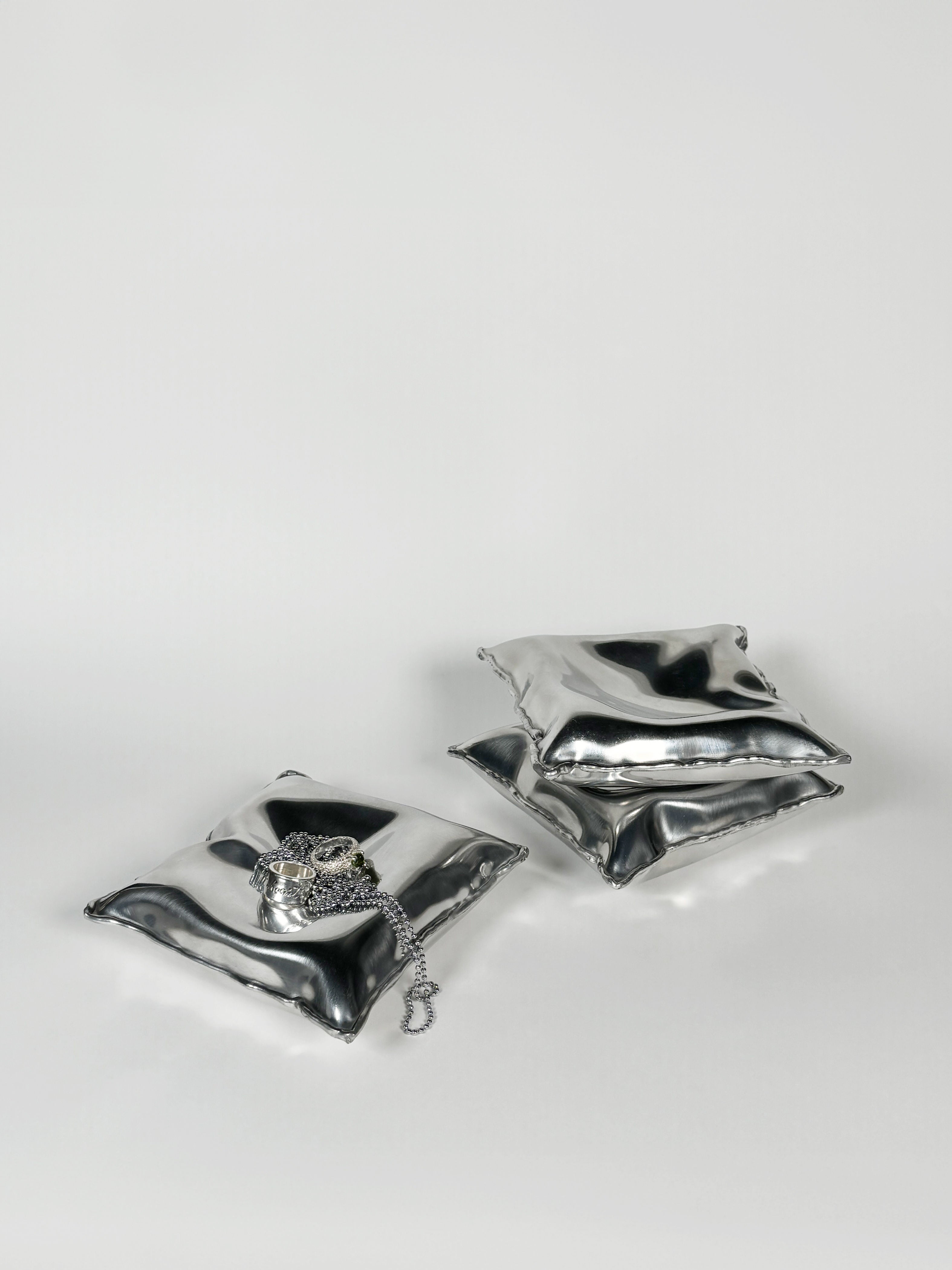 Two shiny, silver pillow-shaped sculptures sit on a smooth surface. One Pillow Bowl Large features a decorative silver chain with a lock draped over it. Handcrafted by Emma Stocklassa in Stockholm, these pieces embrace minimalist design against a plain light grey background.