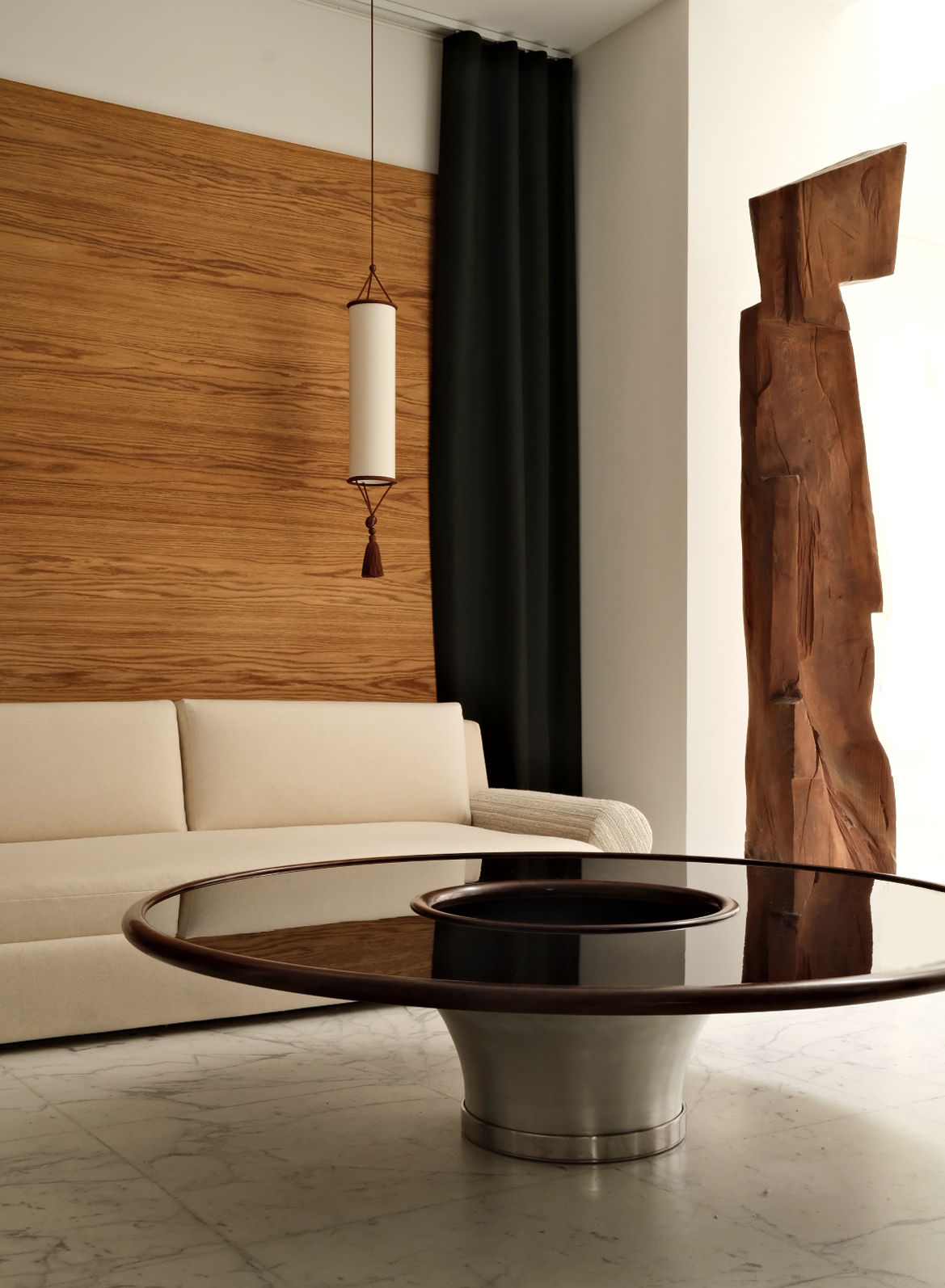 A modern living room showcases a beige sofa against a wooden wall, adorned with dark green curtains, and the "Loulou" Coffee Table by HAUVETTE & MADANI, which features an embossed aluminum finish with a reflective surface. An abstract wooden sculpture enhances the lacquered wood details, while the pristine white marble floor completes the setting.