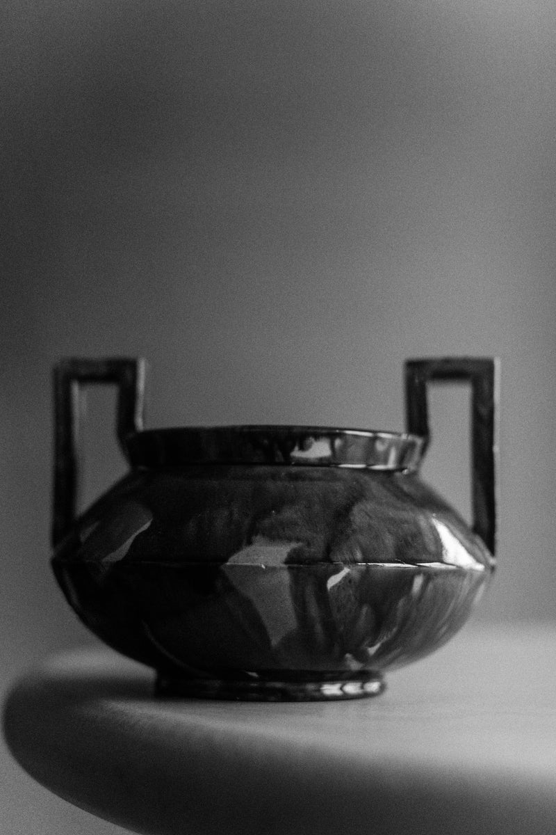 Black and white photo of an antique, metallic Out For Lunch 1930's Höganäs vase with handles, displayed against a soft, blurred background. The focus is on the textured surface and reflective sheen of the vase.
