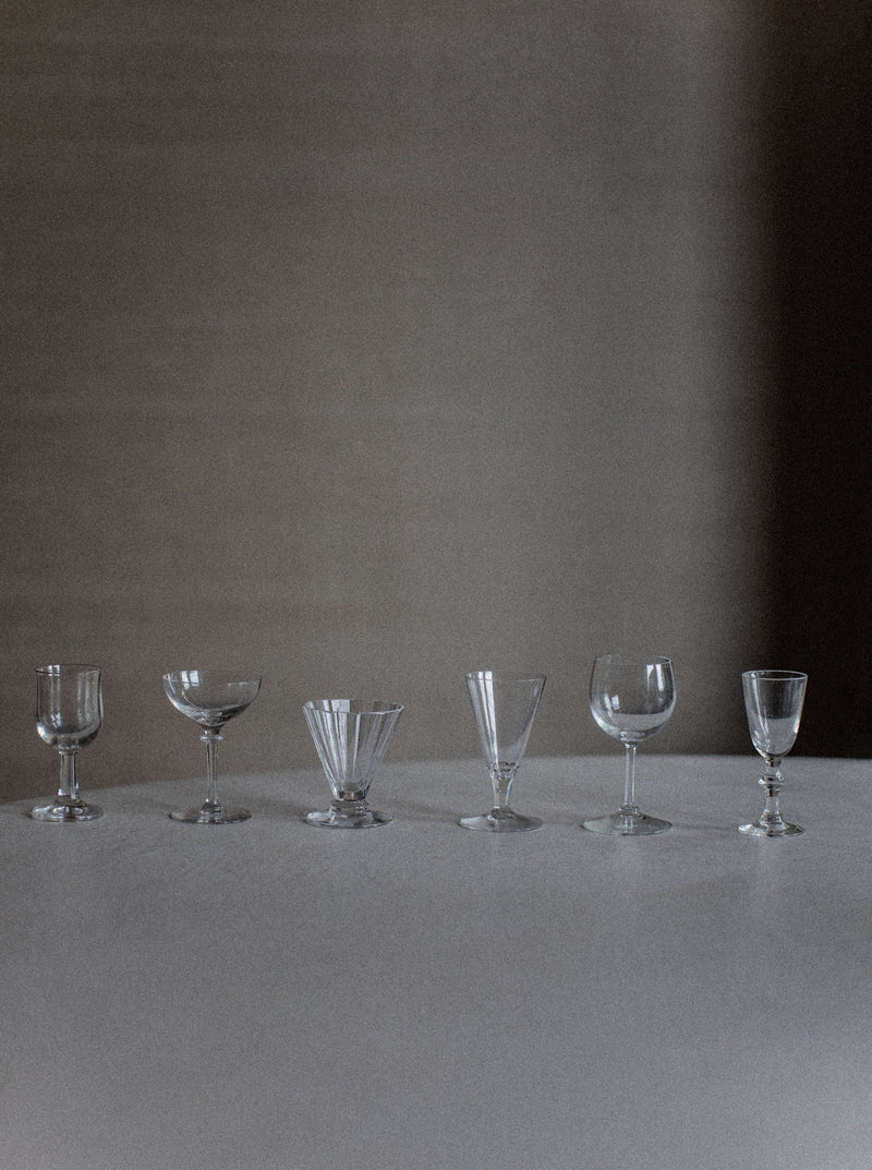 A row of six empty glasses from the "Set of Six Mix-Matched Glasses" by Out For Lunch, including wine glasses, goblets, and cocktail glasses, arranged on a plain surface against a neutral grayish background. The subtle lighting casts shadows that enhance the sophistication and create a serene atmosphere. Each glass narrates its own unique story.