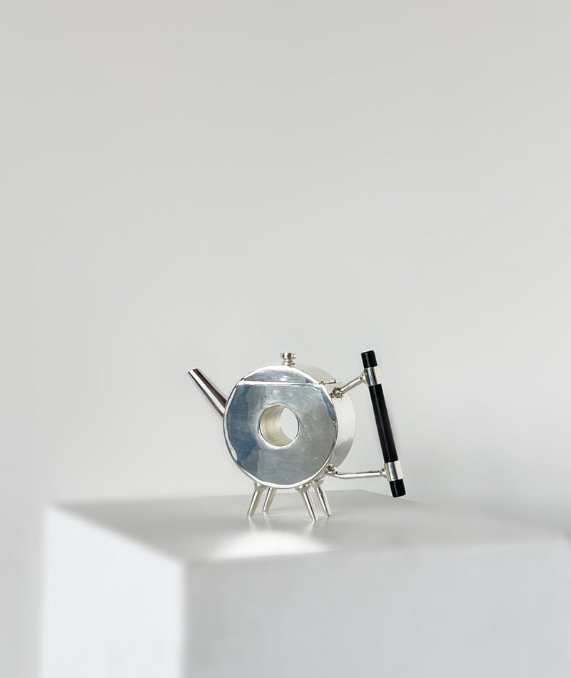 The Christopher Dresser Ornate Round Teapot by Porter Studio is a modern, circular silver-plated teapot with a black handle and angled legs, displayed on a white pedestal against a plain, light gray background.
