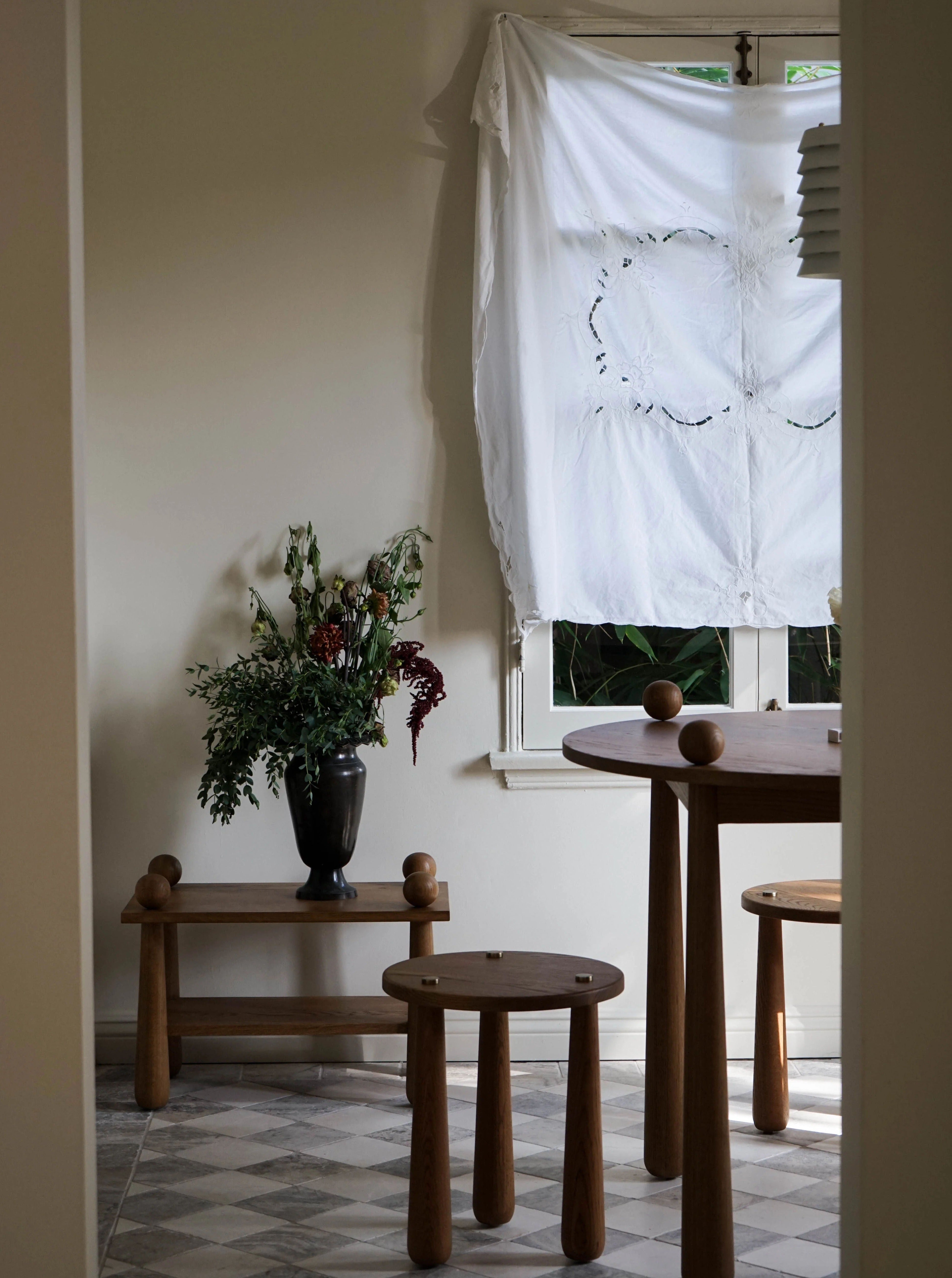 A cozy, minimalist room with a wooden table and Dott Stool, Natural by TASE GALLERY crafted from natural Oak wood sits near a window covered by a sheer white cloth. A small wooden bench holds a black vase filled with an assortment of greenery and flowers. Geometric-patterned tiles cover the floor.