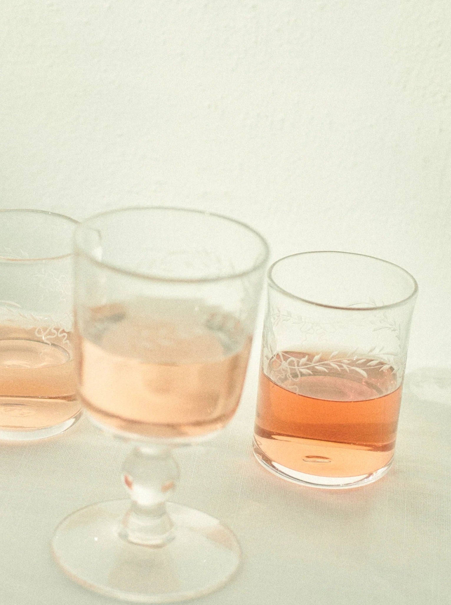 Two glasses with rose wine against a light background, one on a stem and the other a tumbler, both partially full and reflecting soft, warm lighting. The glasses are Akua Objects Barbro Water Glasses - Set of four.