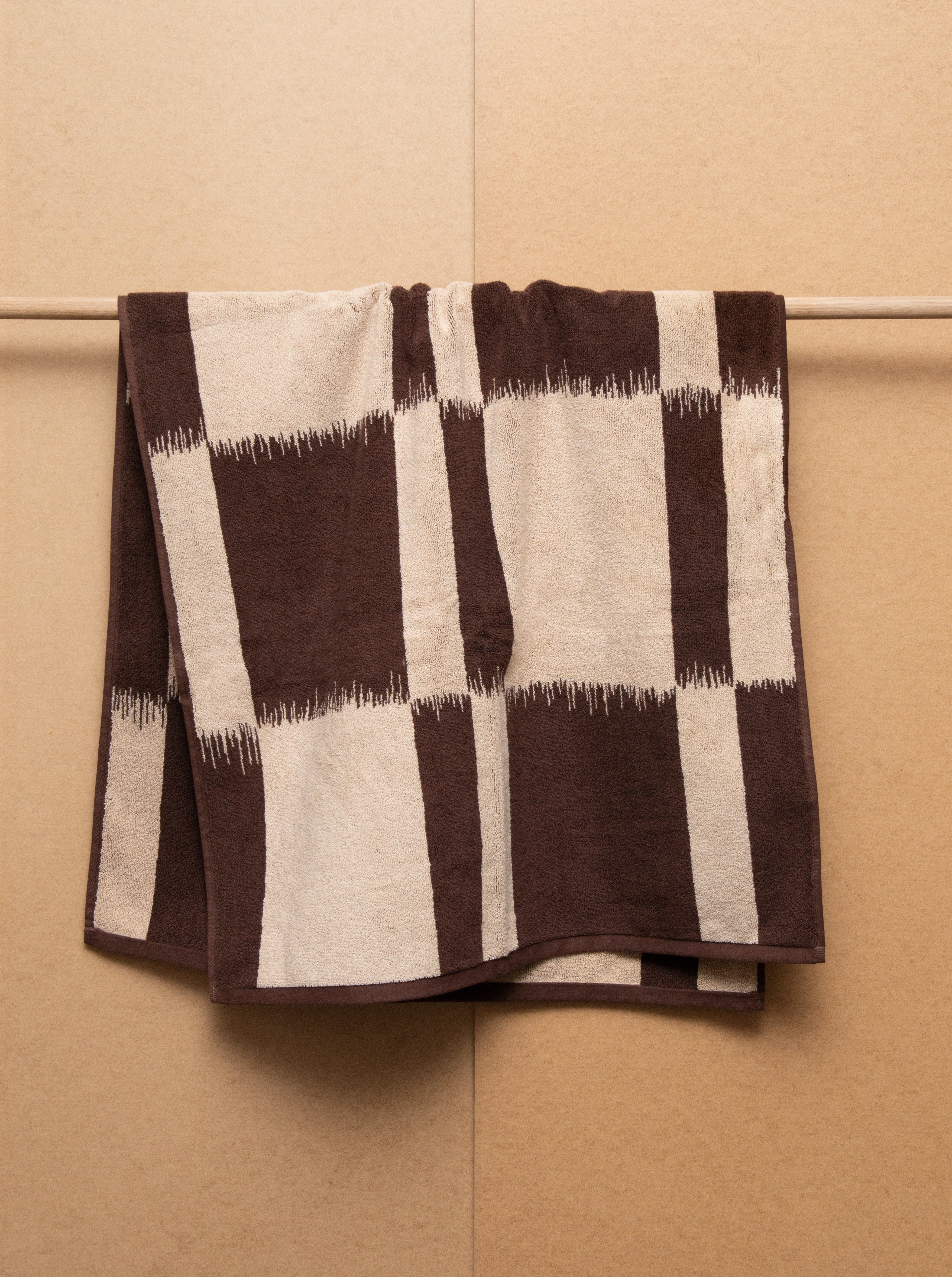 A 600 GSM Karin Bath Towel in brown and beige stripes folded neatly and hung on a horizontal rack against a light brown tiled wall. Brand: Autumn Sonata.