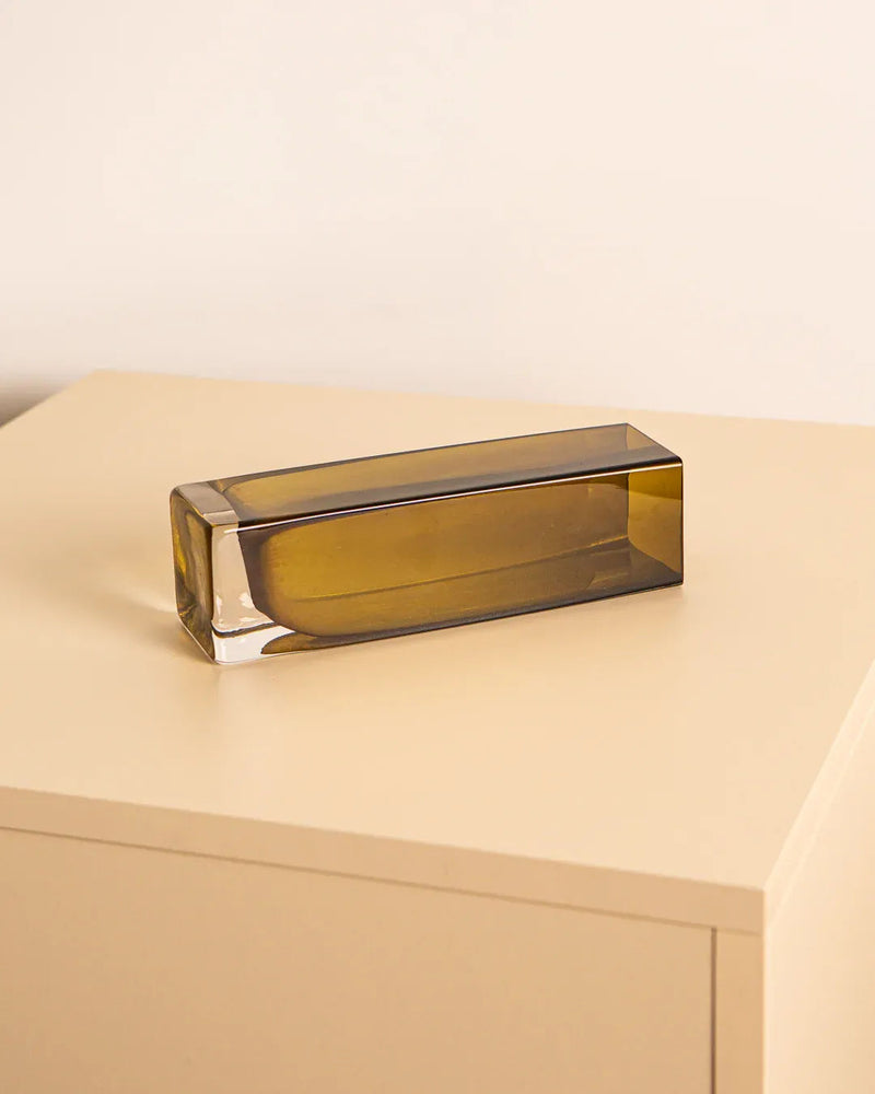 An Amber Murano Glass Vase 80's from Treaptyque rests on a beige surface against a light cream wall. This rectangular, transparent olive-green vase, echoing the elegance of its 1980s design, features smooth, polished edges and a minimalist style, casting a soft shadow beneath it.