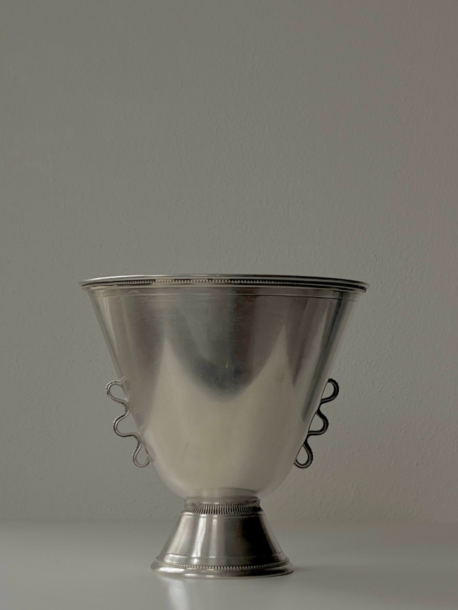 A 1930s Silver Vase by Médecine, featuring intricate handwork and two handles, is elegantly displayed on a sleek pedestal against a plain light gray backdrop. Its reflective surface beautifully captures the minimalist and elegant design, reminiscent of Stockholm's understated charm.