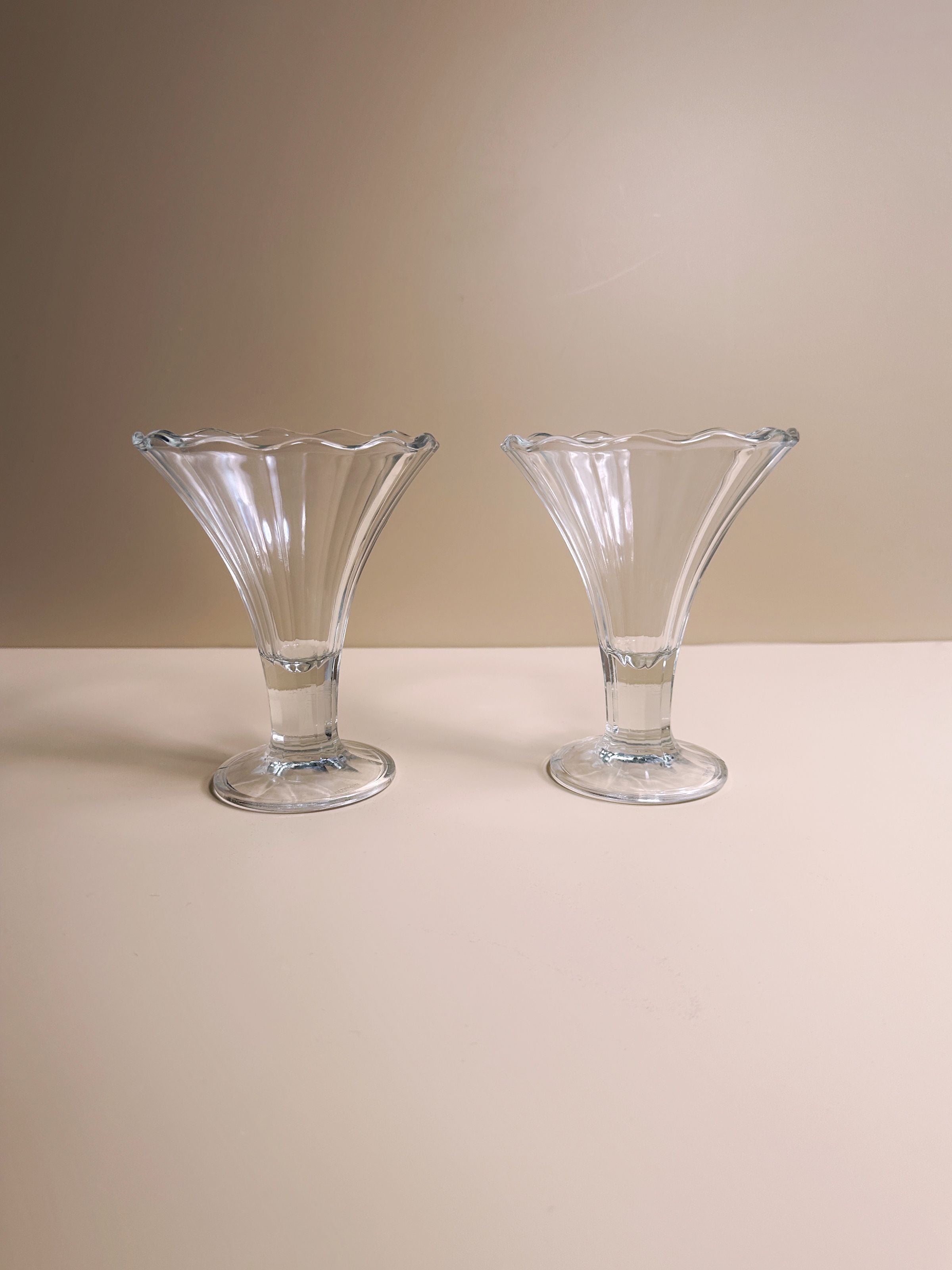 The 2 Italian Art Deco Cocktail Glasses by Dodo Vintage are showcased side by side on a light beige surface against a matching background. With their clear, symmetrical design and fluted edges, these glasses add an elegant touch to any table setting.