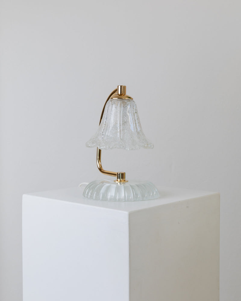 The Bottega Jacobs Murano Glass Table Lamp from 1970s Italy, featuring a vintage glass shade and curved gold-toned stand, rests on a white pedestal against a light gray background. This piece exudes an elegant Hollywood Regency style.