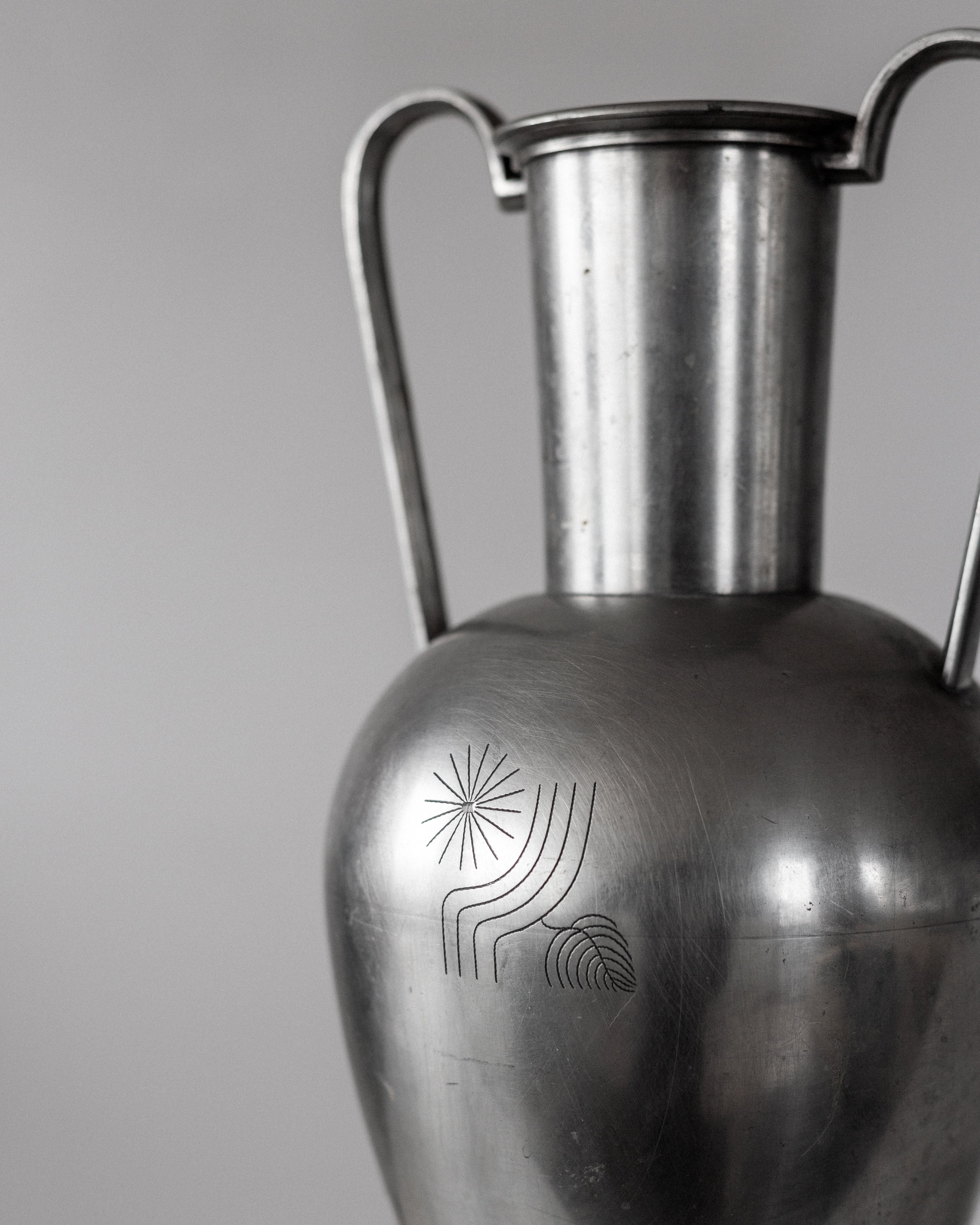 A close-up of the 1930s Spigel Large Modernist Metal Amphora reveals a pewter vase with two handles and a smooth, rounded body. Its engraved abstract design of circular and wavy lines resembles a sun and waves, reflecting Scandinavian metalwork. The background is neutral gray.