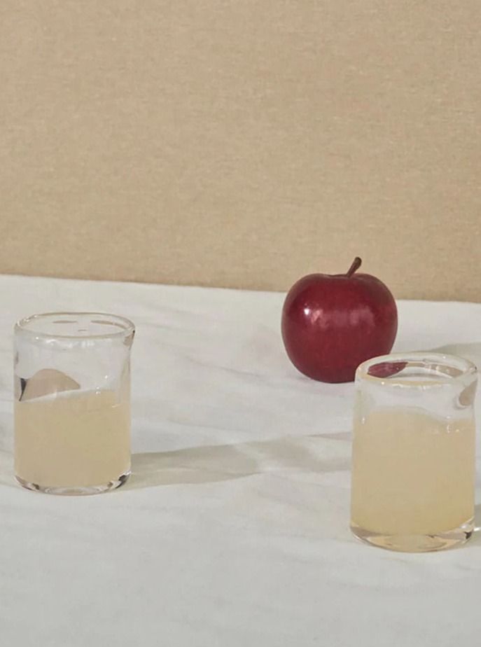 A red apple is placed on a white surface next to two small glasses filled with a pale yellow liquid. The durable, handblown FRAMA 0405 Glass Set of Two | Clear | Small are positioned side by side, slightly closer to the viewer than the apple. The background has a light brown color.