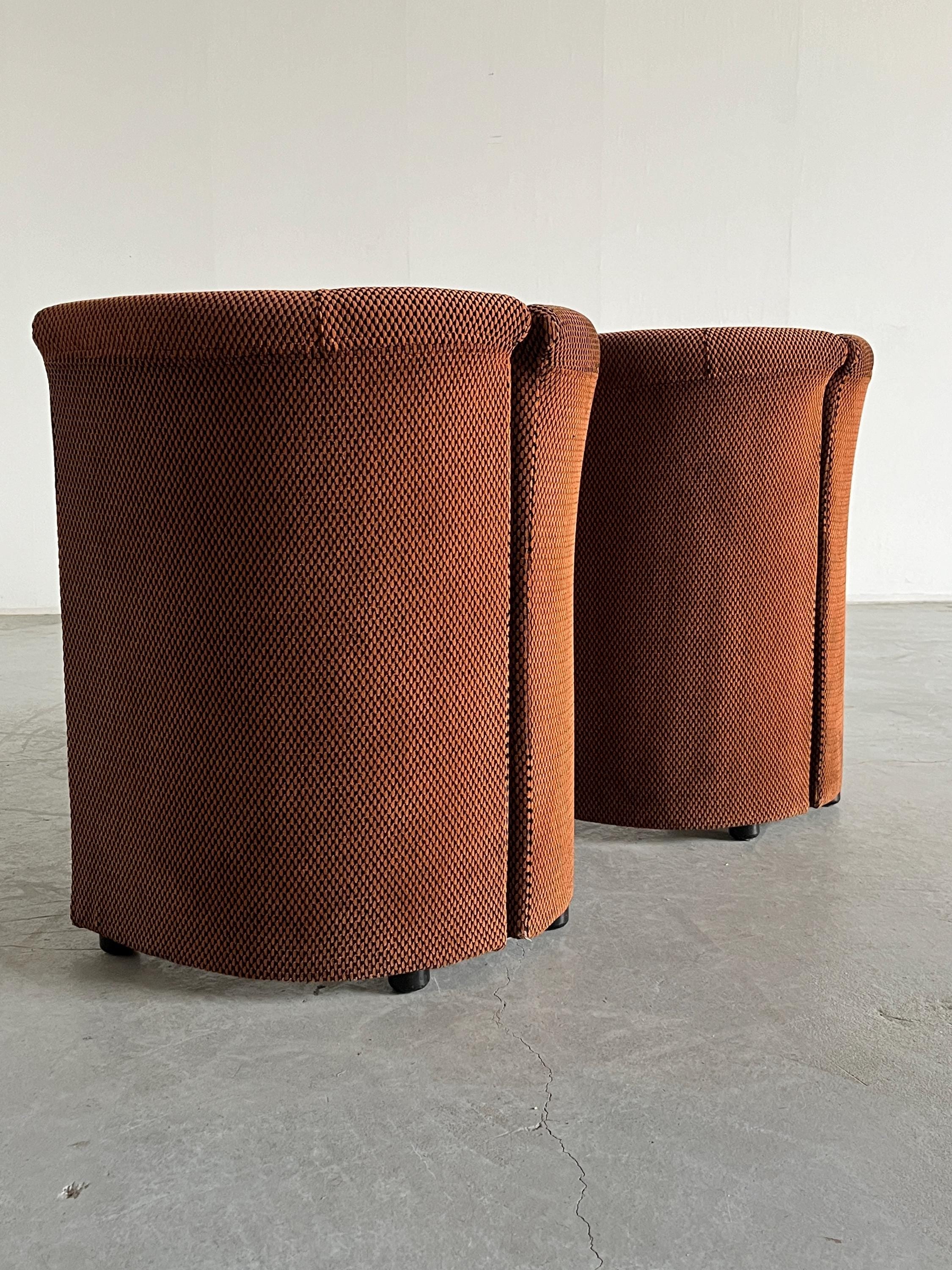 Two rust-colored cylindrical chairs from Cherry Cargo's "Pair of Postmodern Armchairs" collection rest on a light gray concrete floor. Upholstered in textured, checkered fabric and lacking visible armrests or backs, they offer a minimalist, modern design.