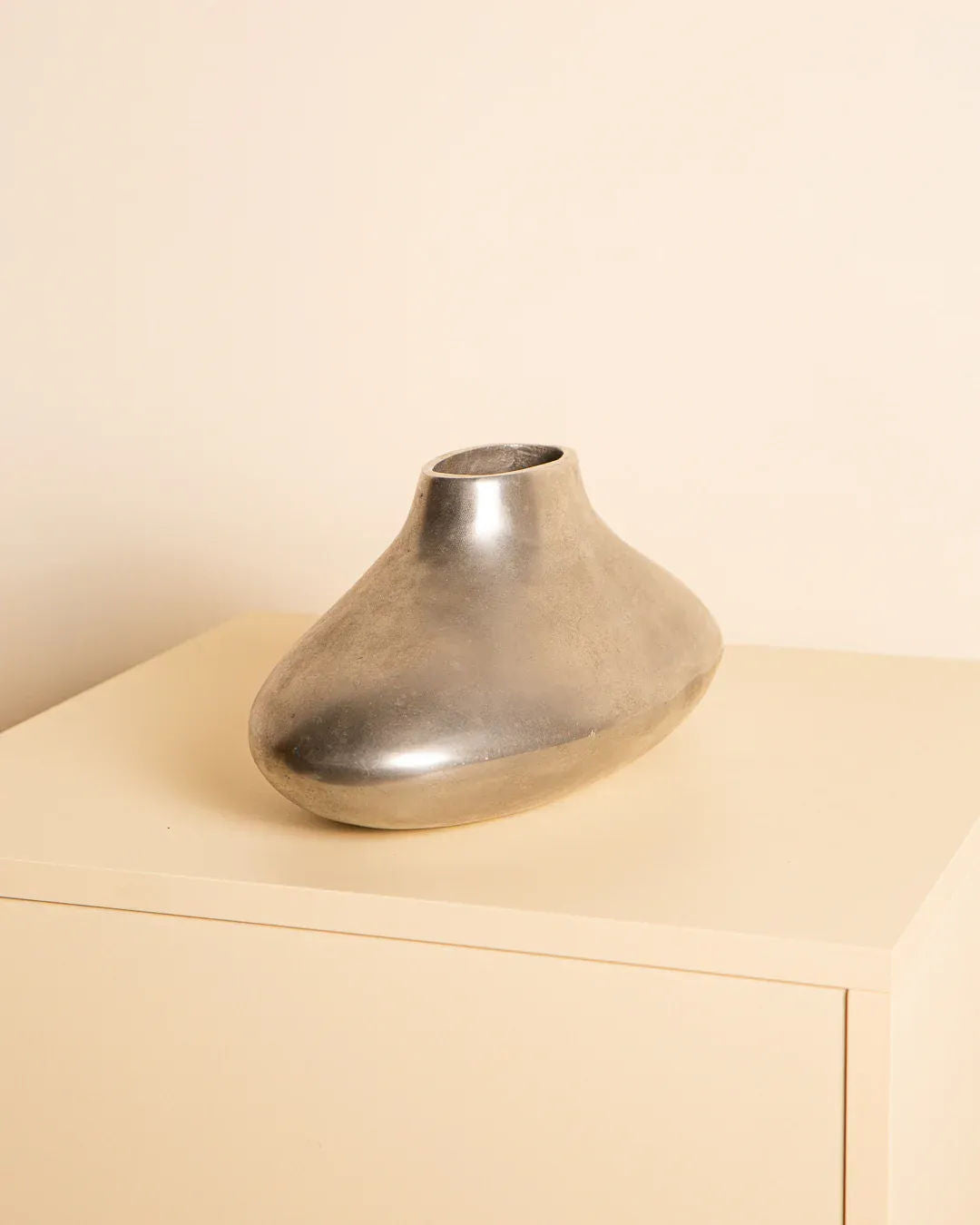 Treaptyque's 80's Cast Aluminium Vase, featuring a smooth metallic finish and natural patina, sits atop a minimalist light-colored table. The Italian-designed vase has an asymmetrical, organic shape with a small top opening against a soft beige background.