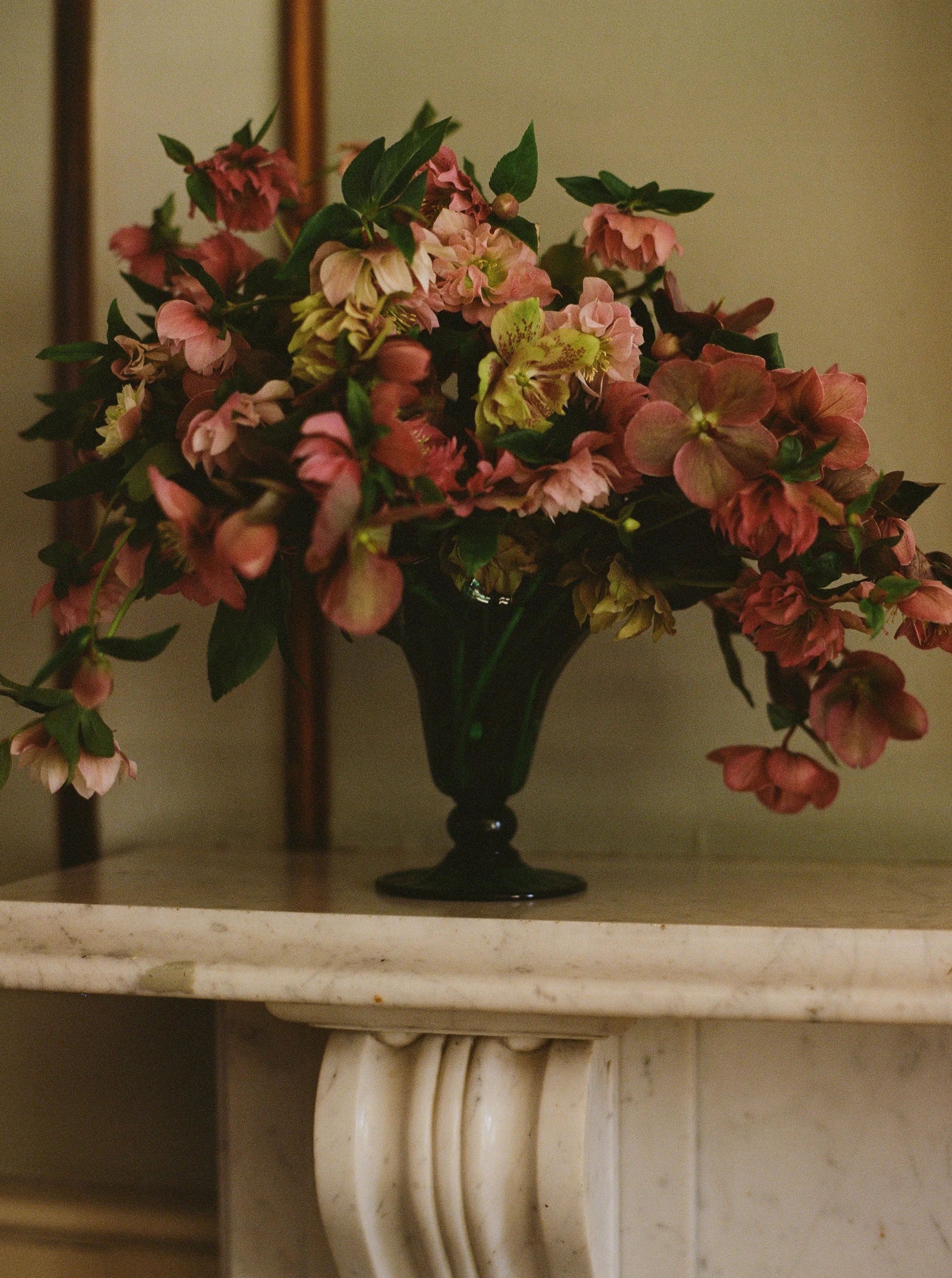 A bouquet of pink flowers with green leaves arranged in a Karen Blixen Vase Small by Akua Objects sits atop a white marble surface. The background features a muted, warm-toned interior setting with hints of wood accents.