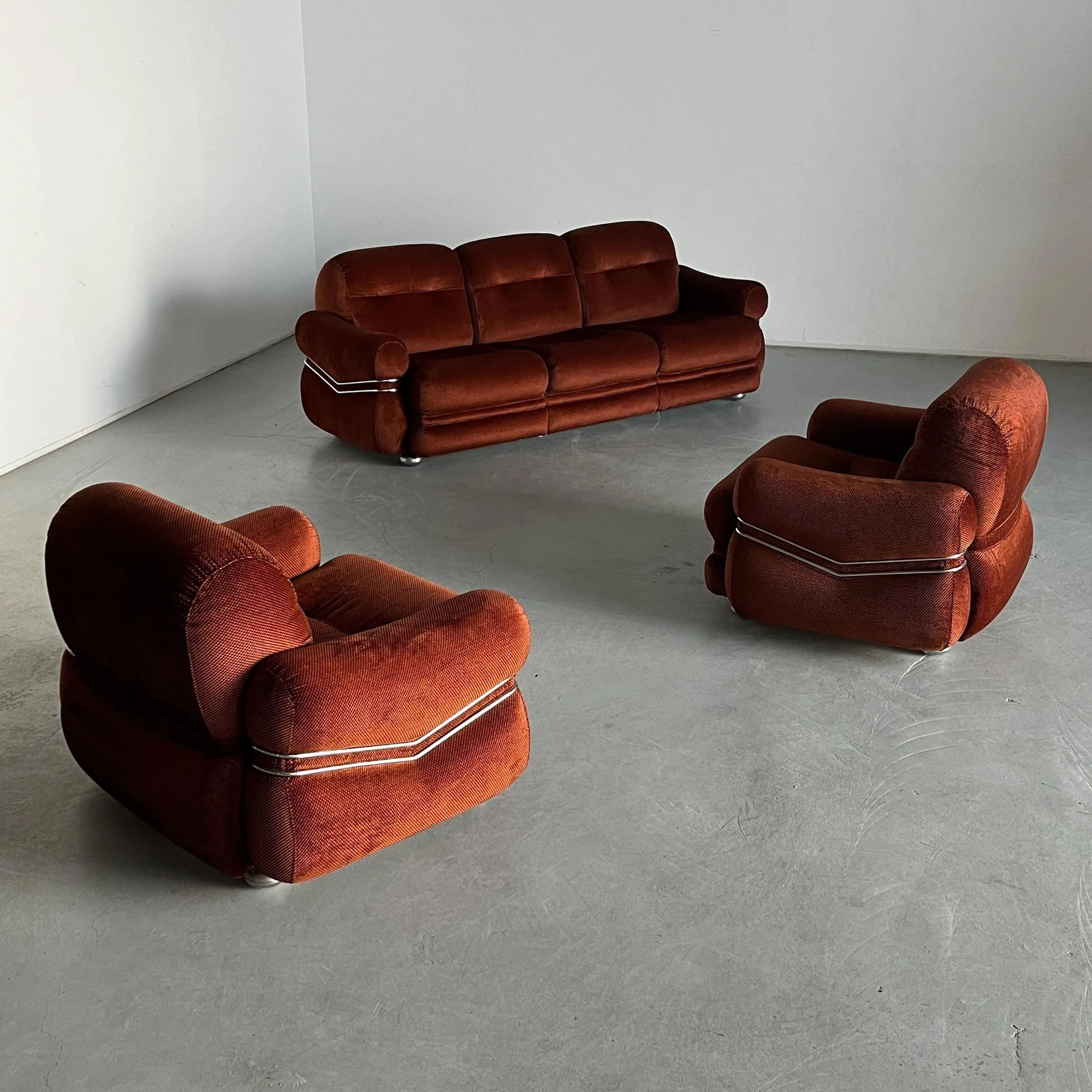 A minimalistic room features a vintage 1970s Italian Space Age seating set by Cherry Cargo, with plush checkered upholstery, rounded edges, and white piping accents. The three-seater sofa and matching armchairs elegantly contrast against the plain white wall and grey floor.