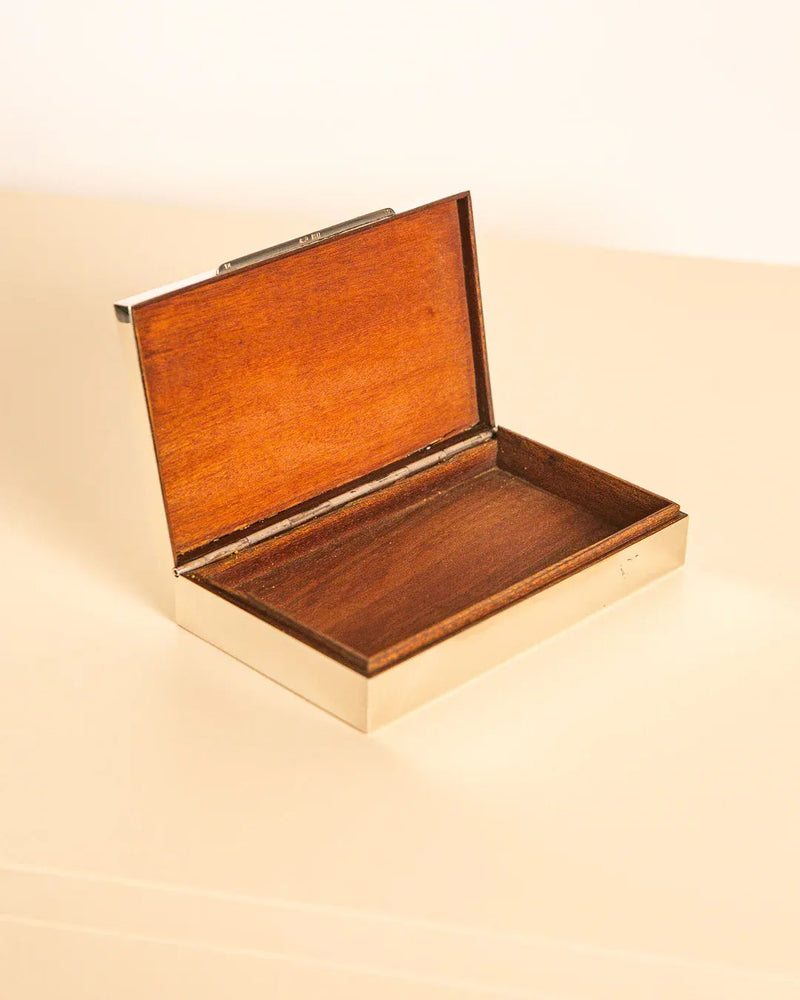 The 60's Silver and Wood Box by Treaptyque, featuring a sleek silver metal frame and an open wooden design inspired by refined 1960s Italian aesthetics, rests gracefully on a cream-colored surface. The interior of the box is empty, and the lid opens with elegant hinges.