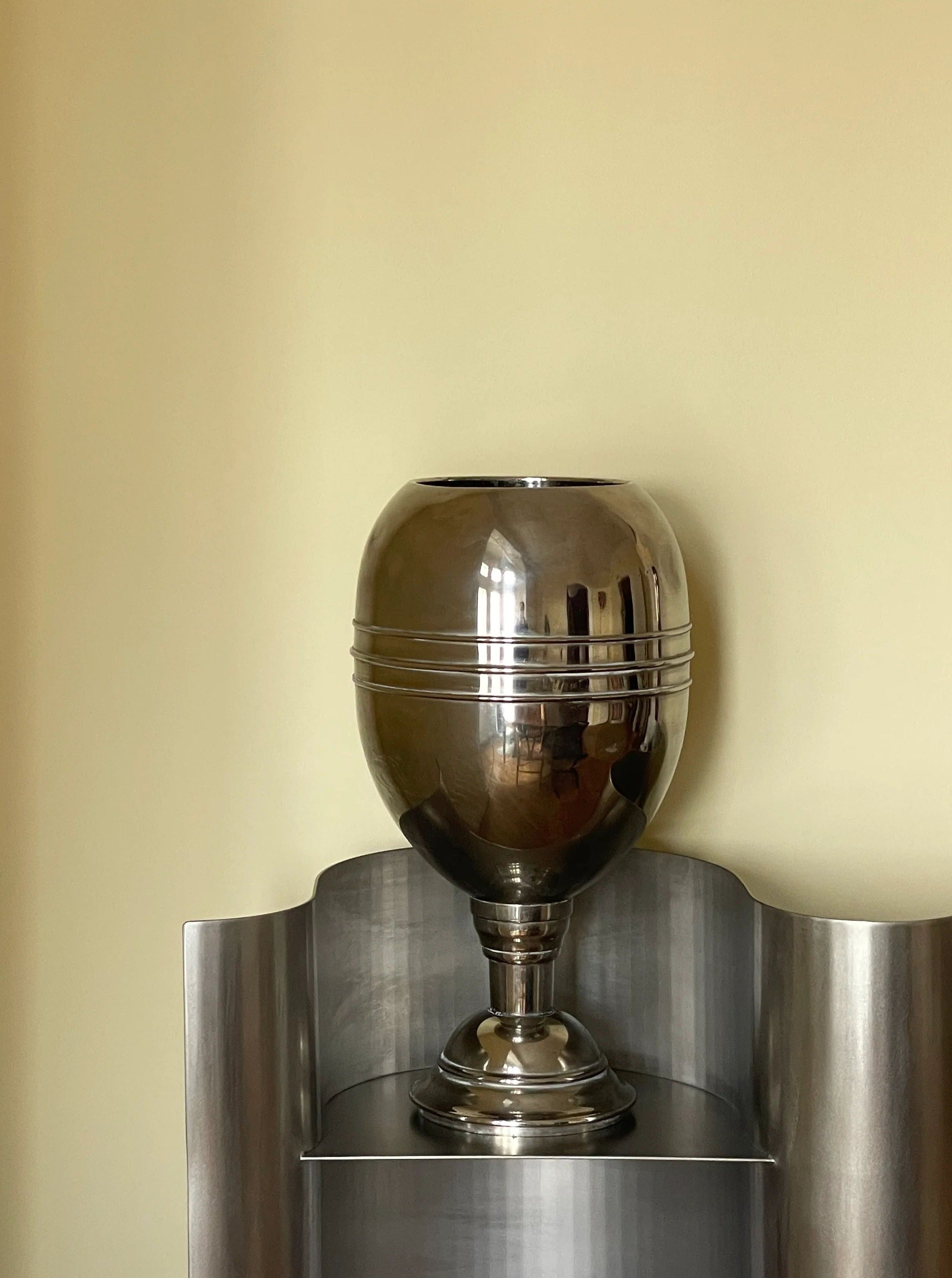 A sleek, steel Big steel vase with a reflective surface sits on a minimalistic pedestal against a plain light yellow wall. The trophy is shaped like a globe on a narrow stand. (Collect Cph)