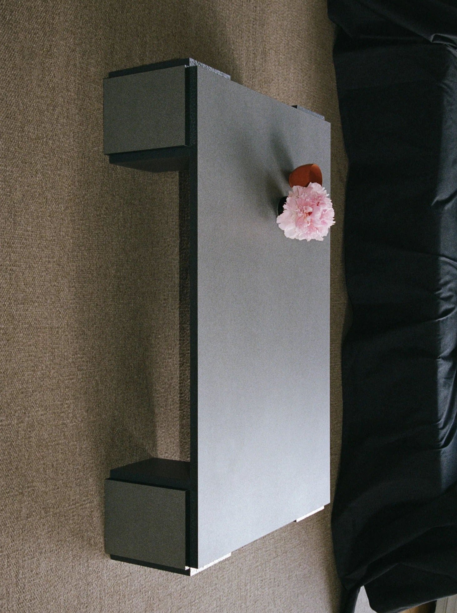 The "Mara" Coffee Table – Night Noise from TEGET, with its minimalist design and dark surface, supports a small vase containing a pink flower atop a neutral-colored carpet. Nearby, there is a piece of dark fabric draped across the side of this elegantly laminated furniture, possibly serving as a blanket or sofa cover.