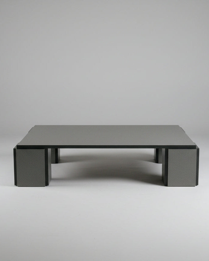 The "Mara" Coffee Table – Night Noise from TEGET, with its minimalist design and dark surface, supports a small vase containing a pink flower atop a neutral-colored carpet. Nearby, there is a piece of dark fabric draped across the side of this elegantly laminated furniture, possibly serving as a blanket or sofa cover.