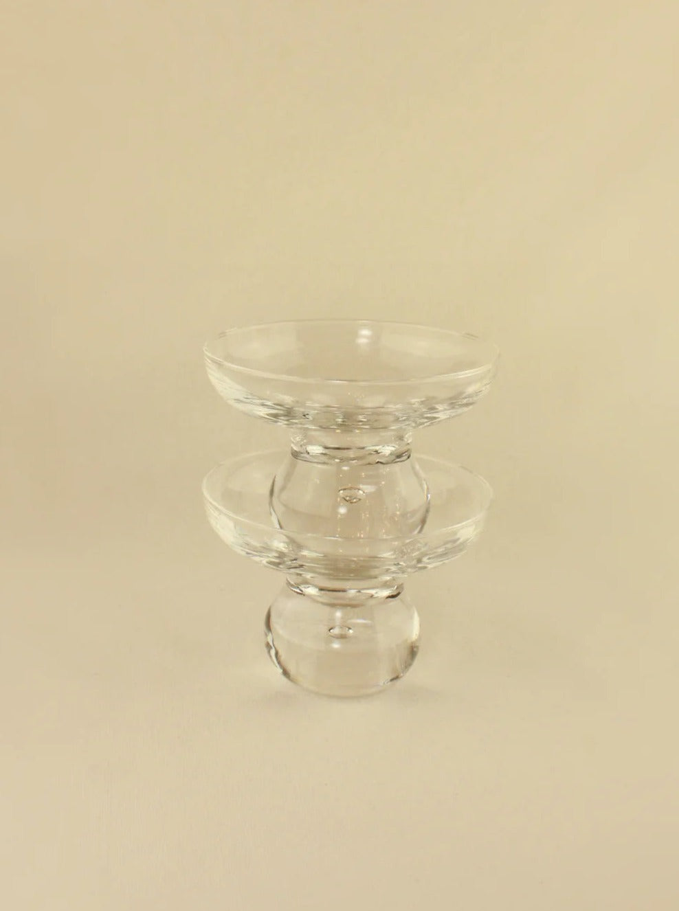 A clear glass candle holder from Boga Avante Shop stands on a neutral background, reminiscent of vintage Italian Glasses. It features an hourglass shape with a wide, flat base for stability, a narrow center, and a slightly flared top. The holder's transparent design catches and reflects light beautifully.