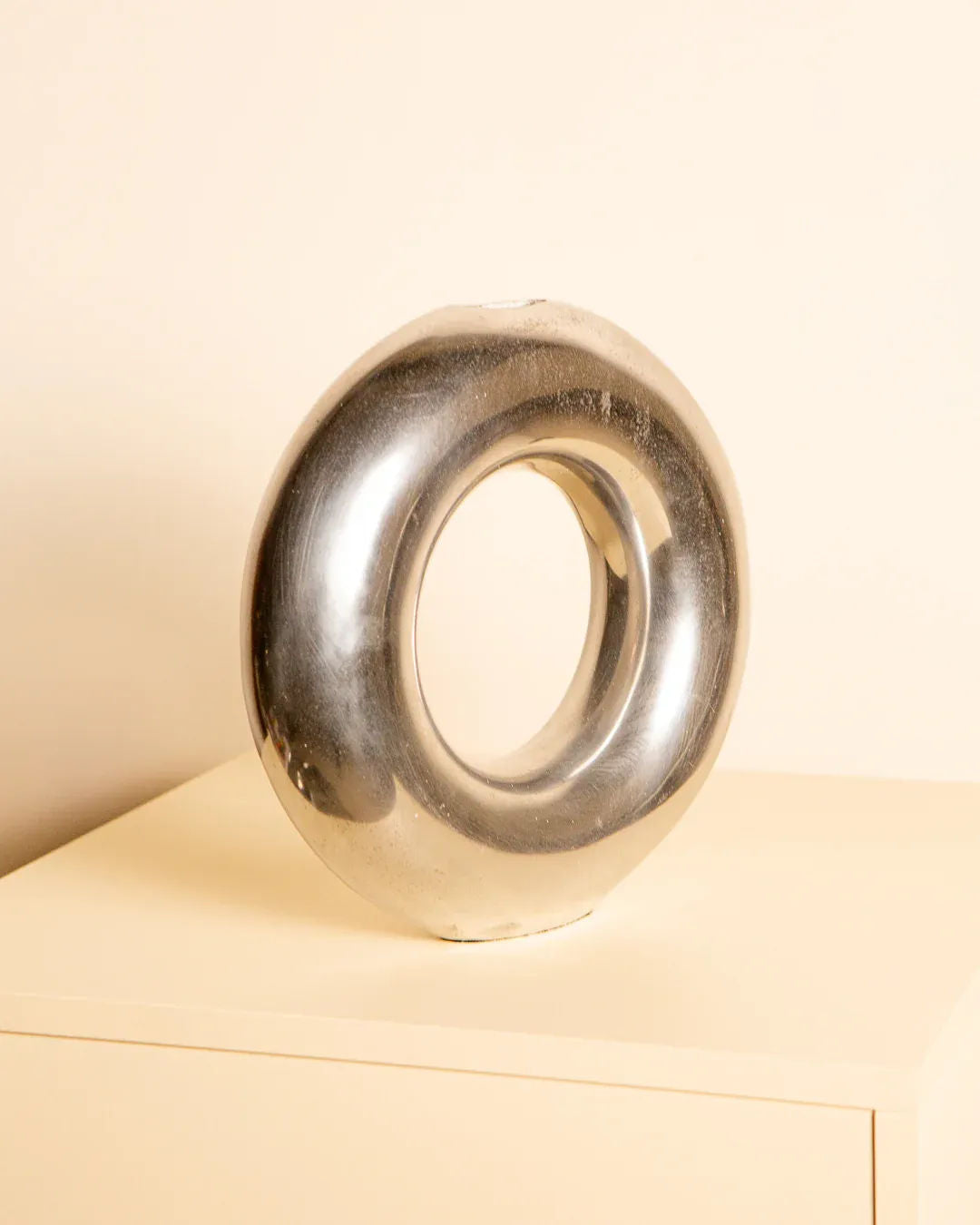 The Treaptyque 80's Aluminum “Circle” Vase, an Italian modern metallic donut-shaped piece, sits on a light surface against a beige background.