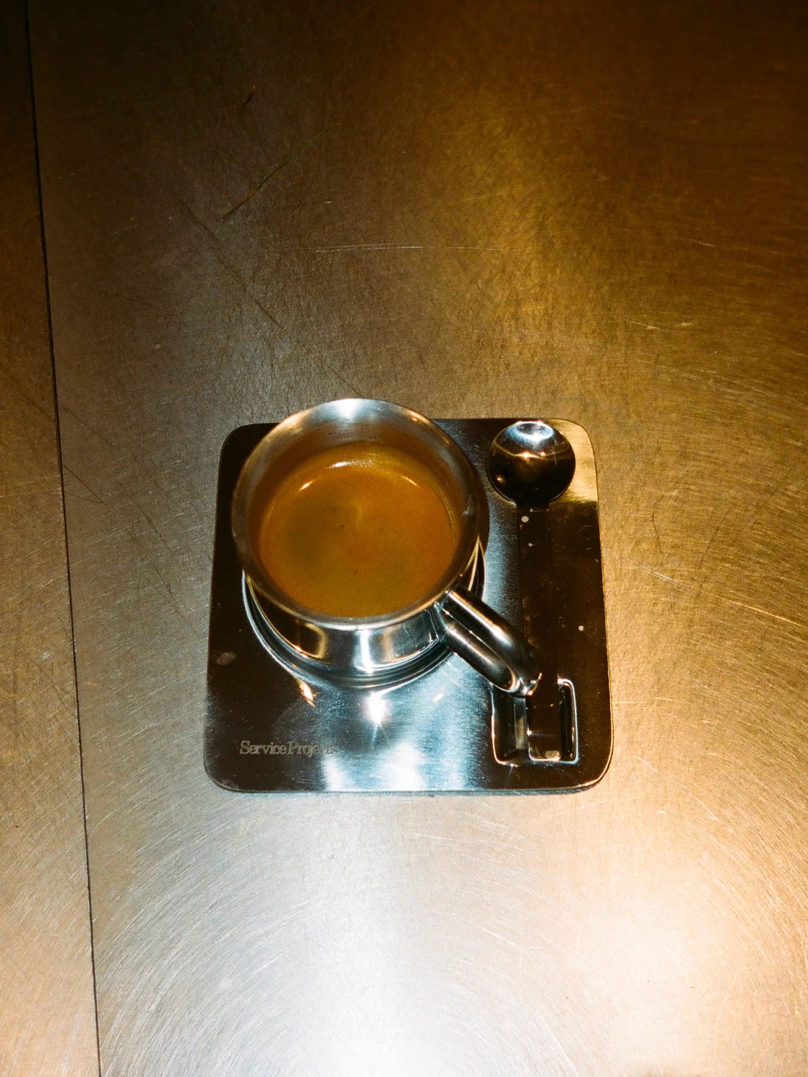 A single espresso in a metal cup with a handle sits on The Arlo Espresso set tray by Service Projects. Reflecting elegant Italian design, the square stainless steel tray holds a small metal spoon beside the cup, and rests on a shiny metal surface.