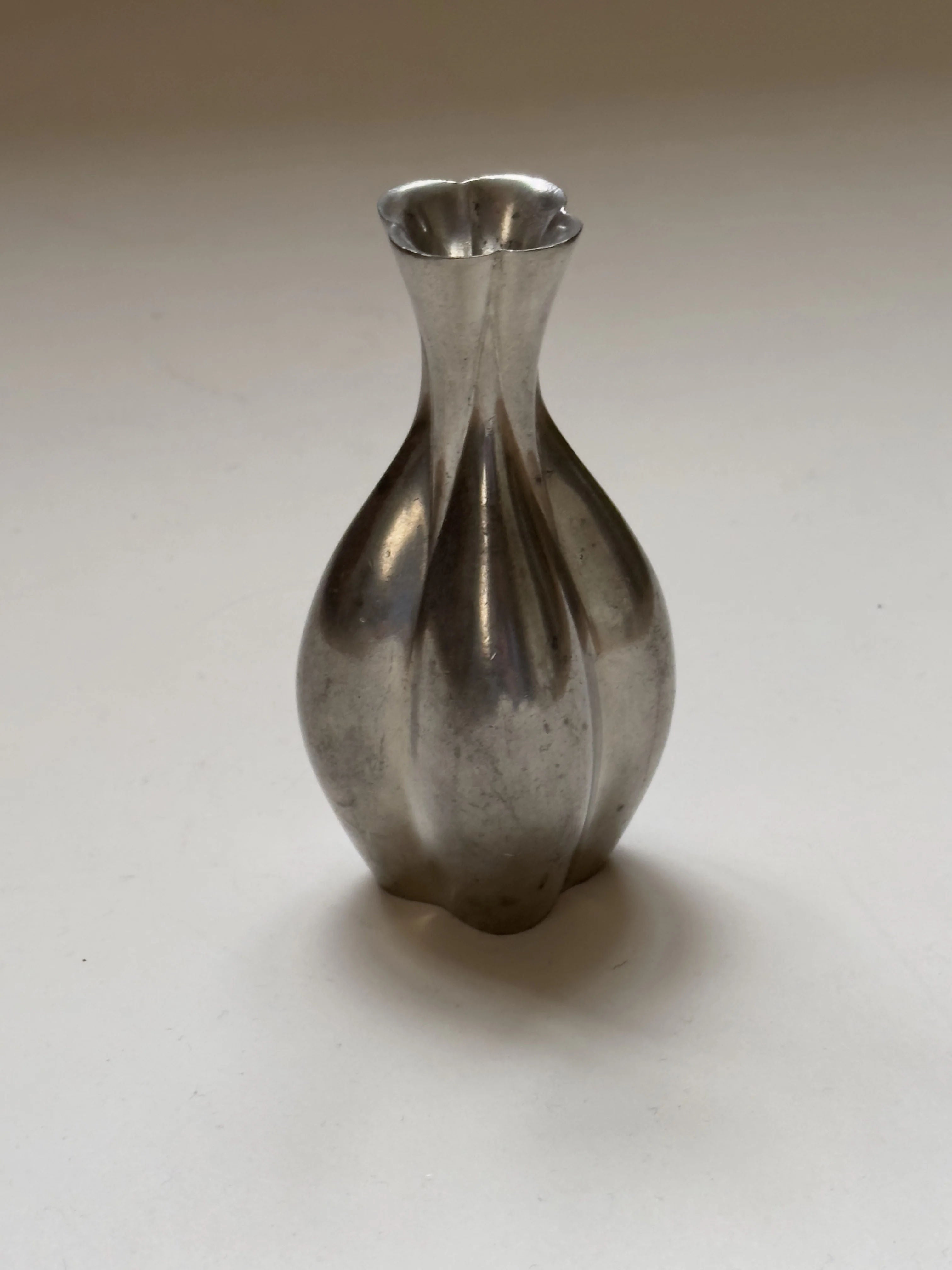 A small, metallic Just Andersen Pewter Vase from Galerie Storm, showcasing a curvy bulbous shape and narrow neck, sits on a plain light-colored surface. This vintage piece features a reflective, silver-like finish.