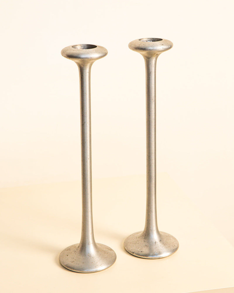 The Treaptyque Pair of Cast Aluminum Candleholders from the '80s features a minimalist industrial design with tall, slender forms, wide round bases, and flared rims, displayed side by side on a light surface against a neutral backdrop.