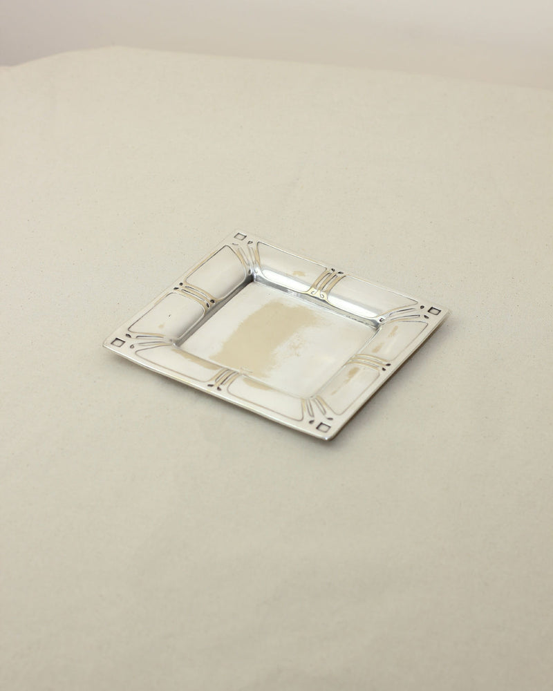 The Silver Modernist Vide-poches from Boga Avante Shop is a small, square silver tray with decorative edges, a shiny reflective finish, and minimalistic design, displayed on a plain light-colored surface.
